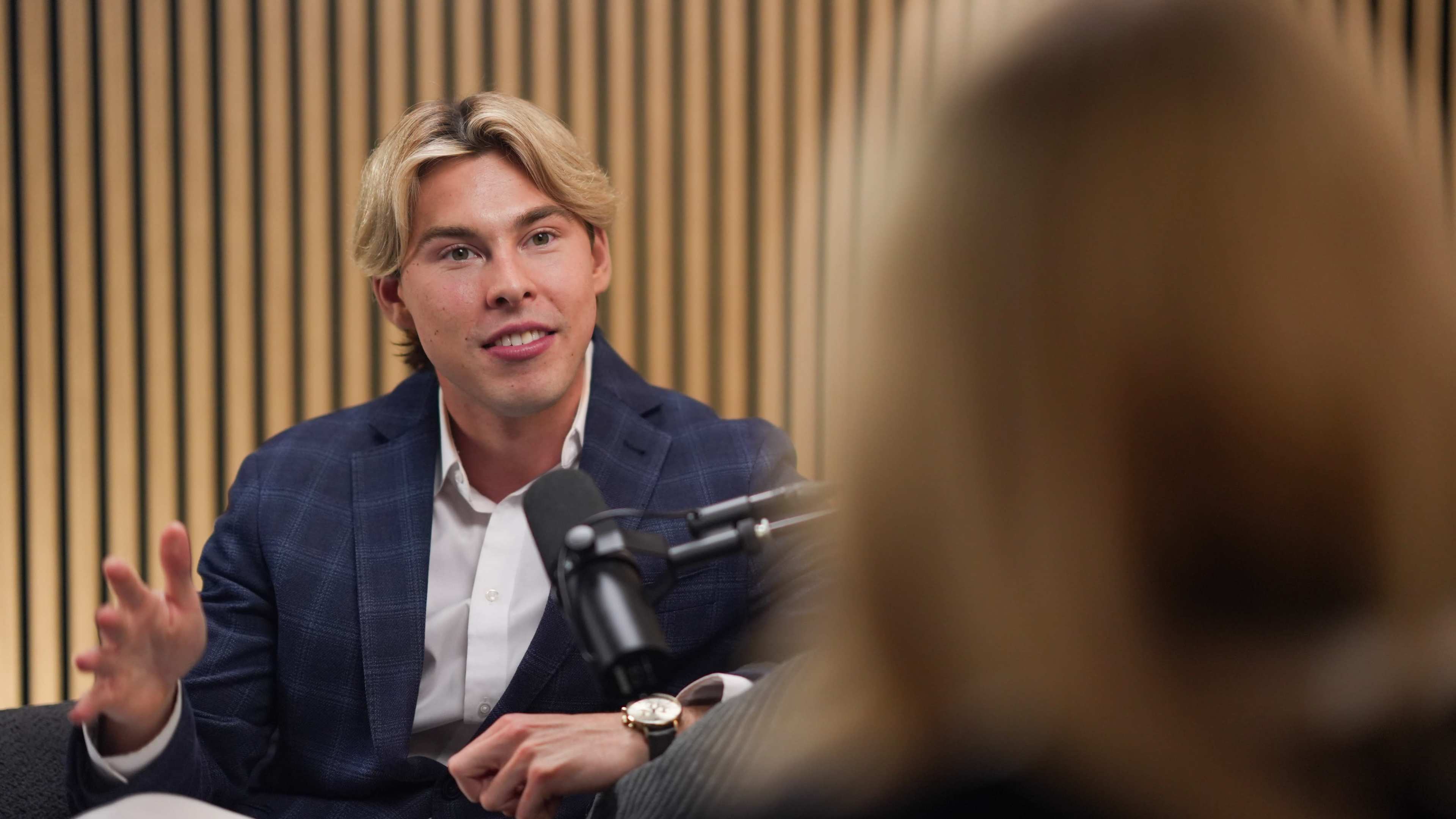 A young man with blonde hair is seated and speaking during a podcast recording, facing a woman out of frame, in a room with wooden paneling.