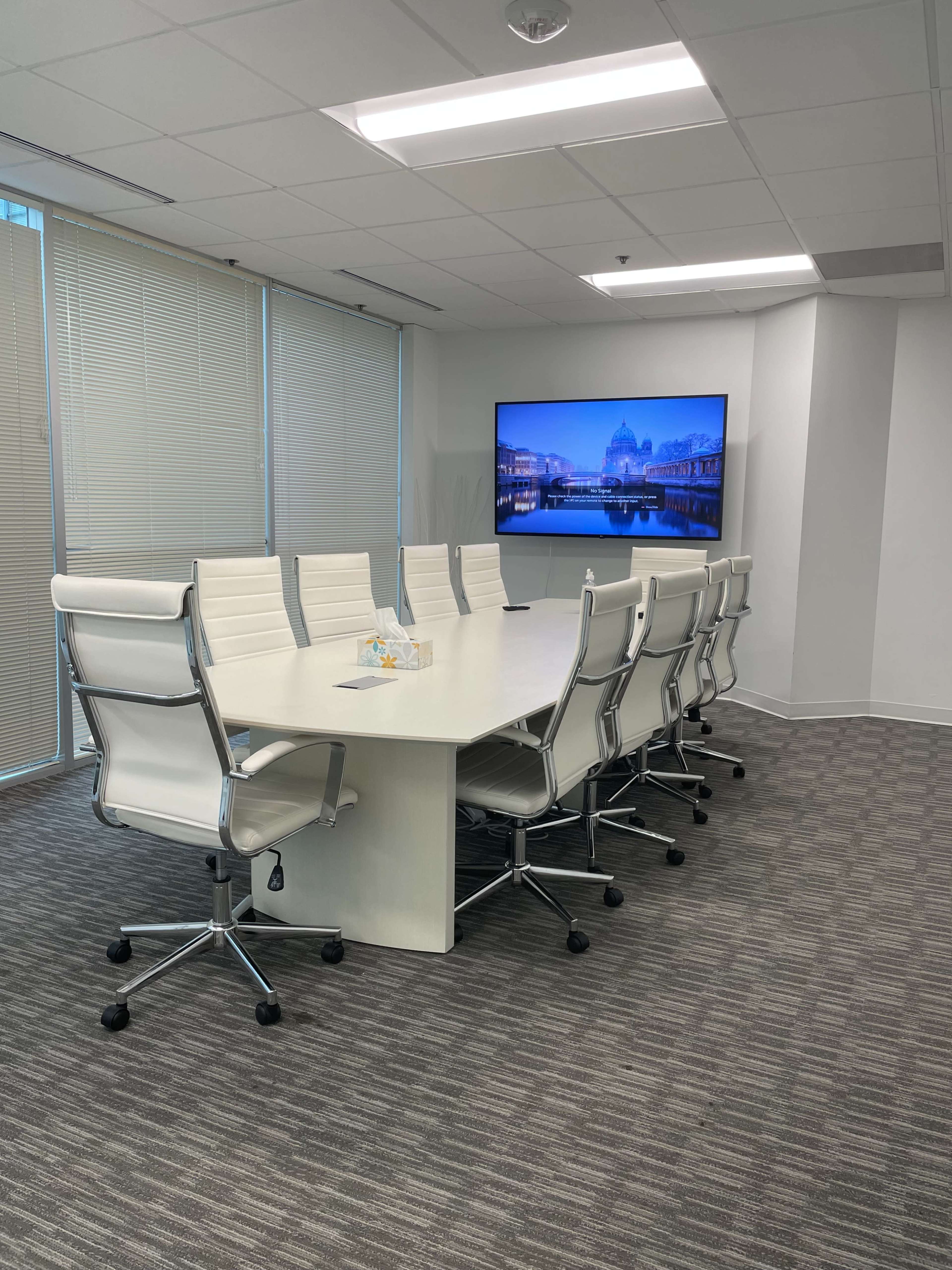 A modern conference room features a long white table surrounded by ergonomic chairs and a large wall-mounted screen displaying a cityscape.
