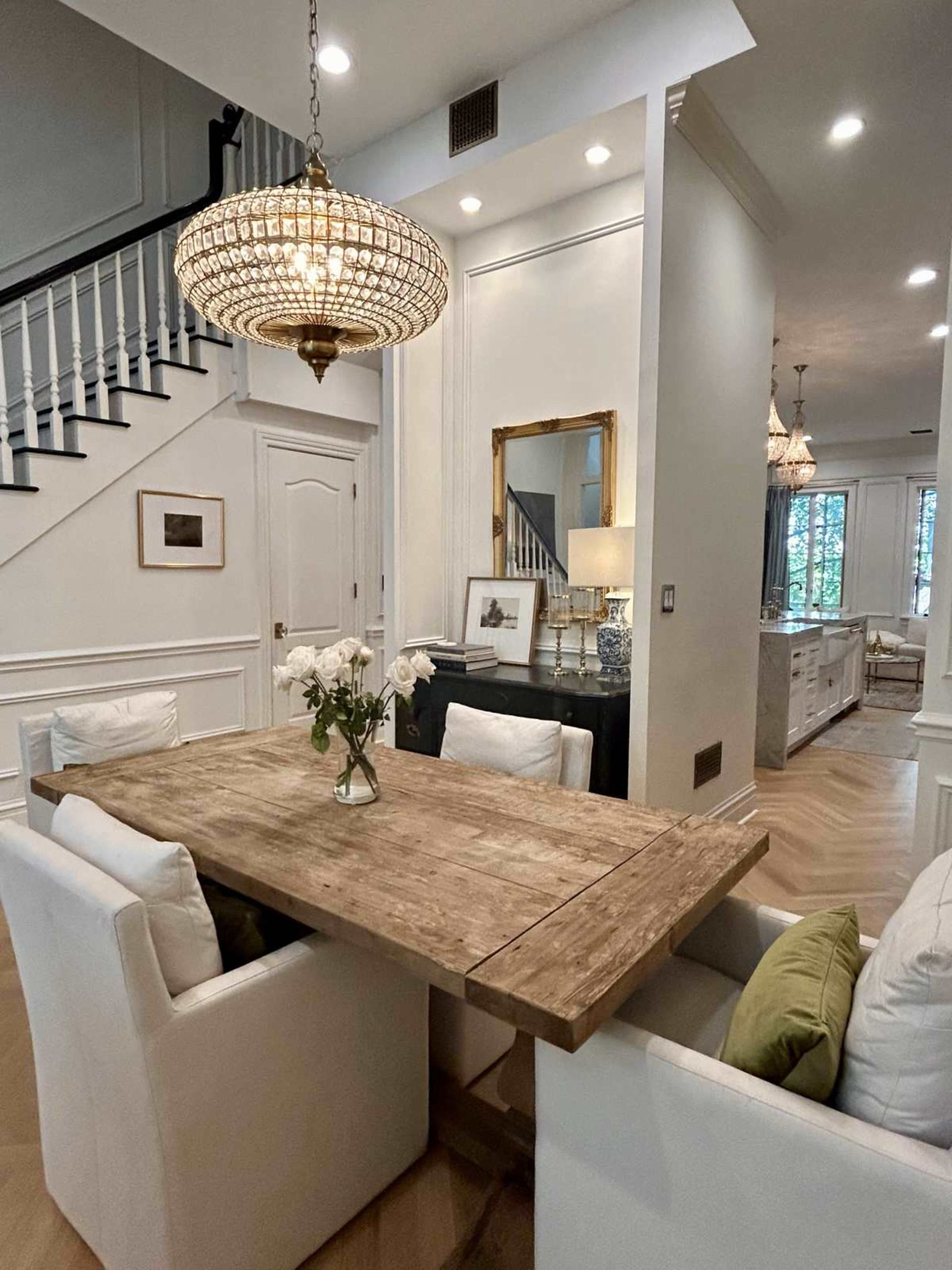 A wooden dining table with white chairs is positioned near a staircase in a well-lit dining area, featuring a chandelier and decorative mirror.