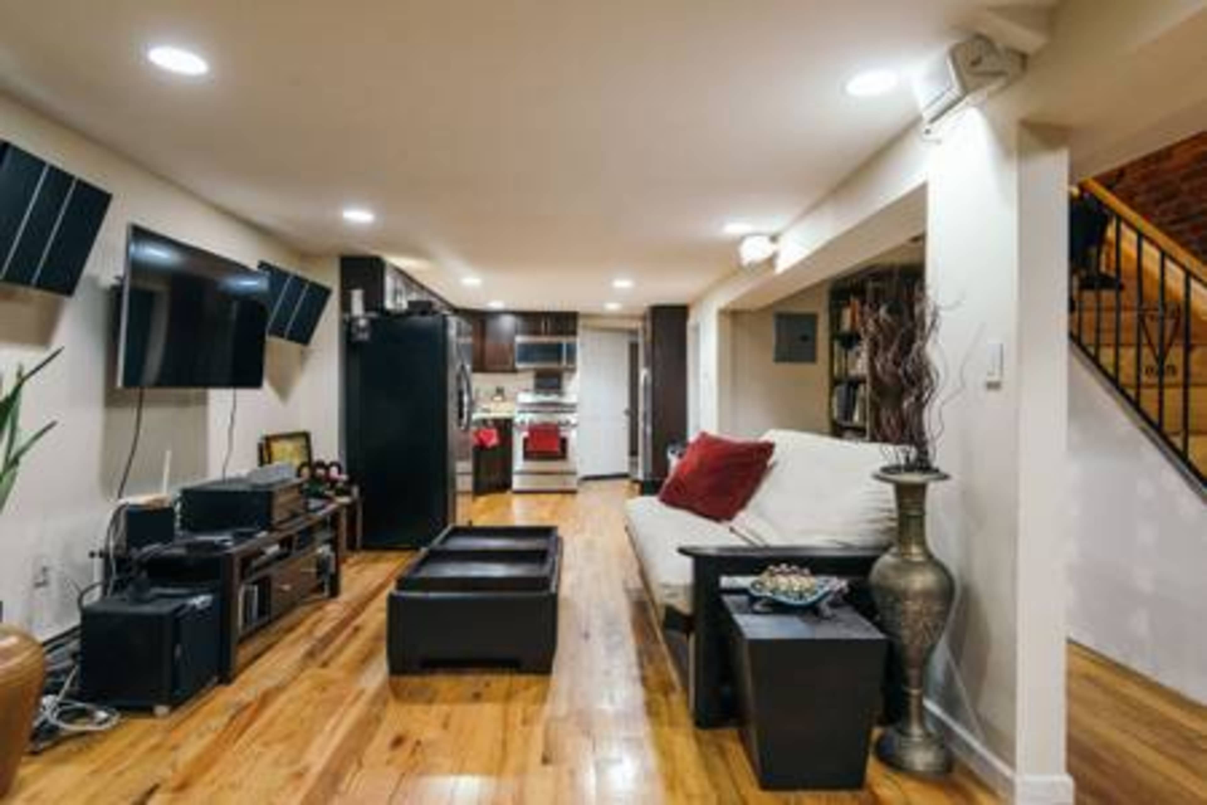 A modern living room features a sectional sofa, a coffee table, and a kitchen area with stainless steel appliances, alongside wall-mounted televisions.