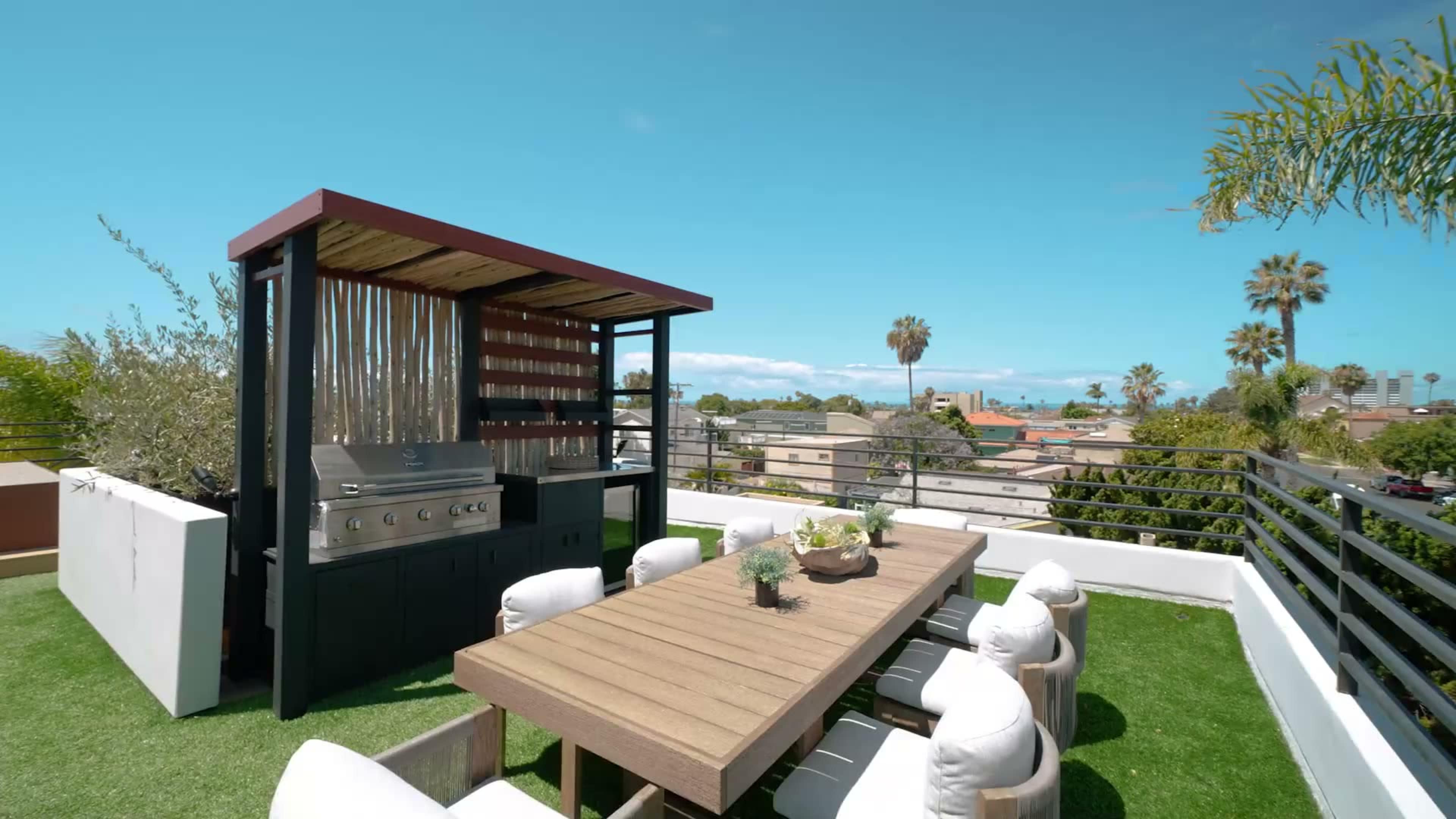 The image shows a rooftop outdoor dining area featuring a wooden table, cushioned chairs, and a barbecue grill under a shaded structure, with a clear blue sky and cityscape in the background.