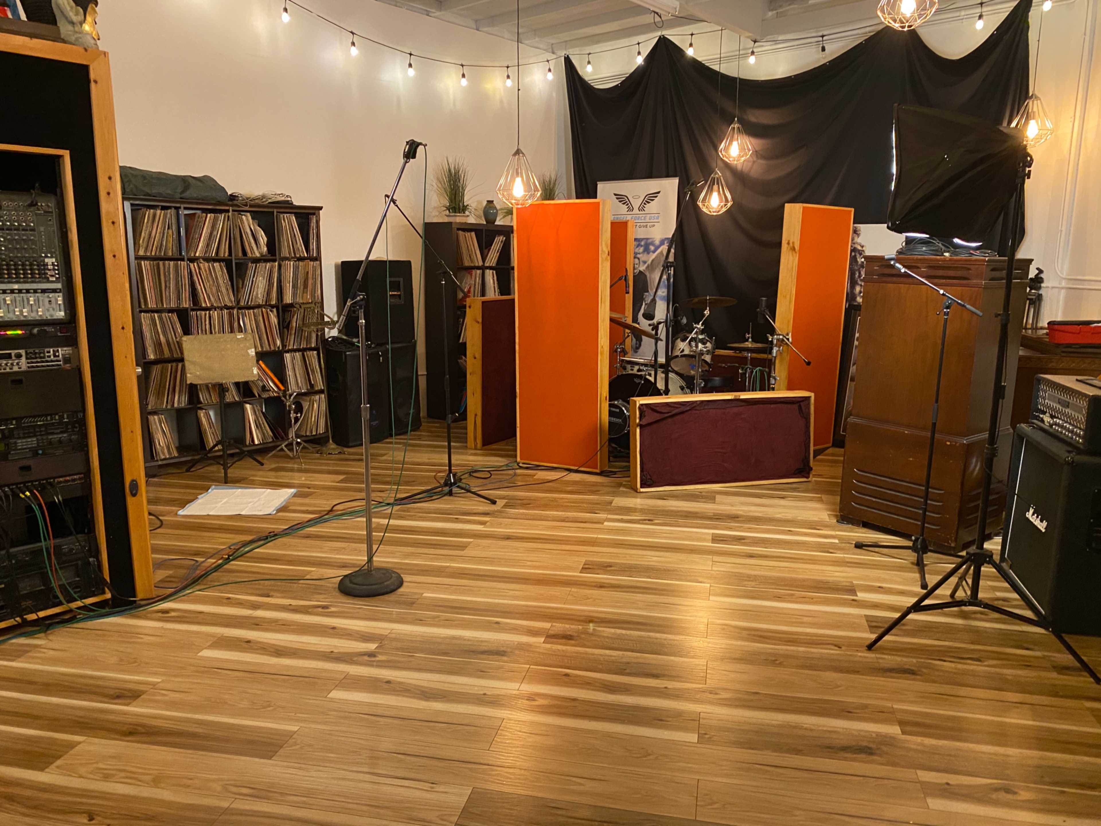 A music studio with wooden flooring, vinyl records on shelves, and a drum set behind soundproof panels.