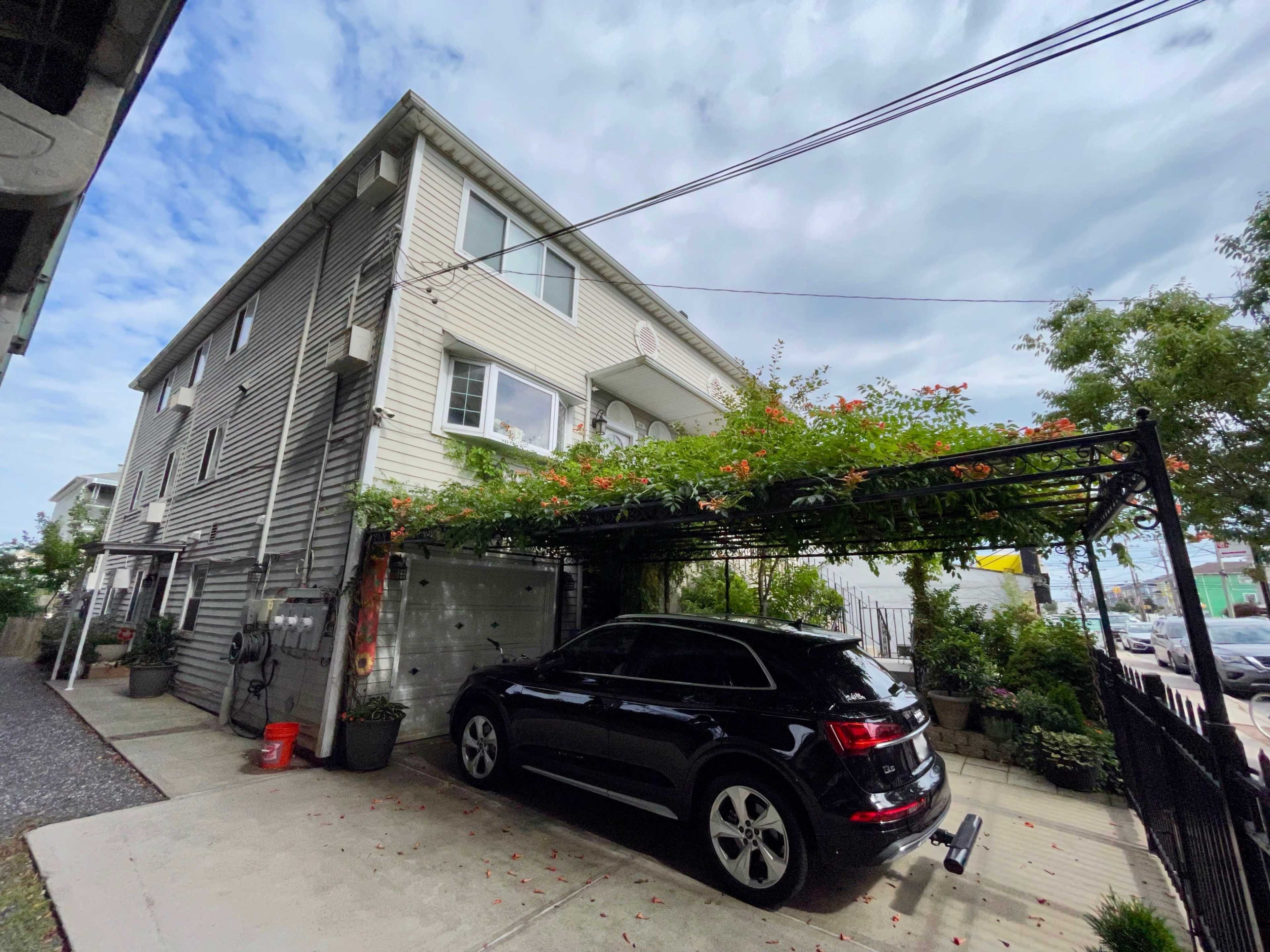 A black vehicle is parked under a carport covered with greenery next to a multi-story residential building.