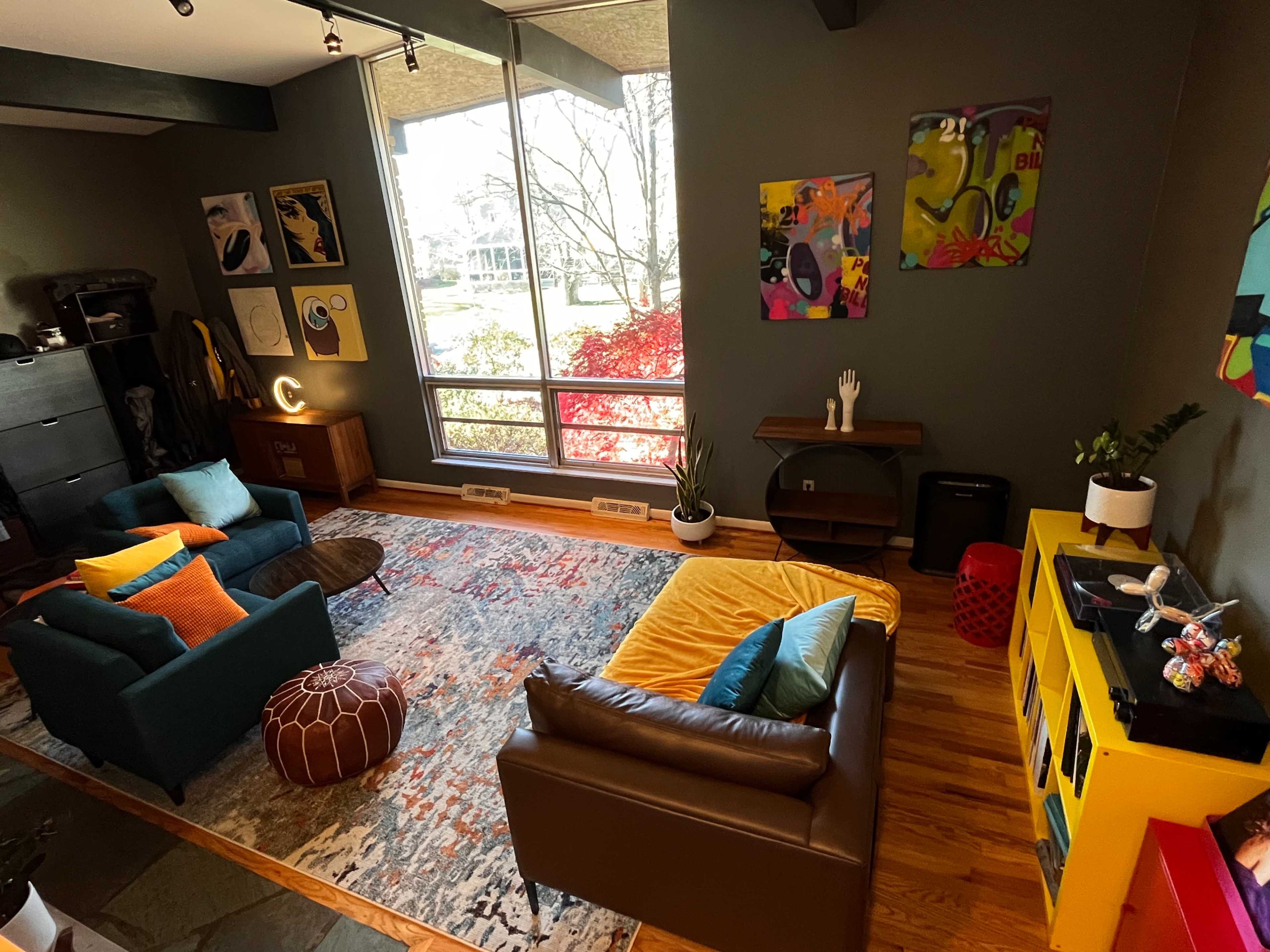 A modern living room with a combination of blue and brown sofas, a colorful rug, and large windows overlooking a garden.