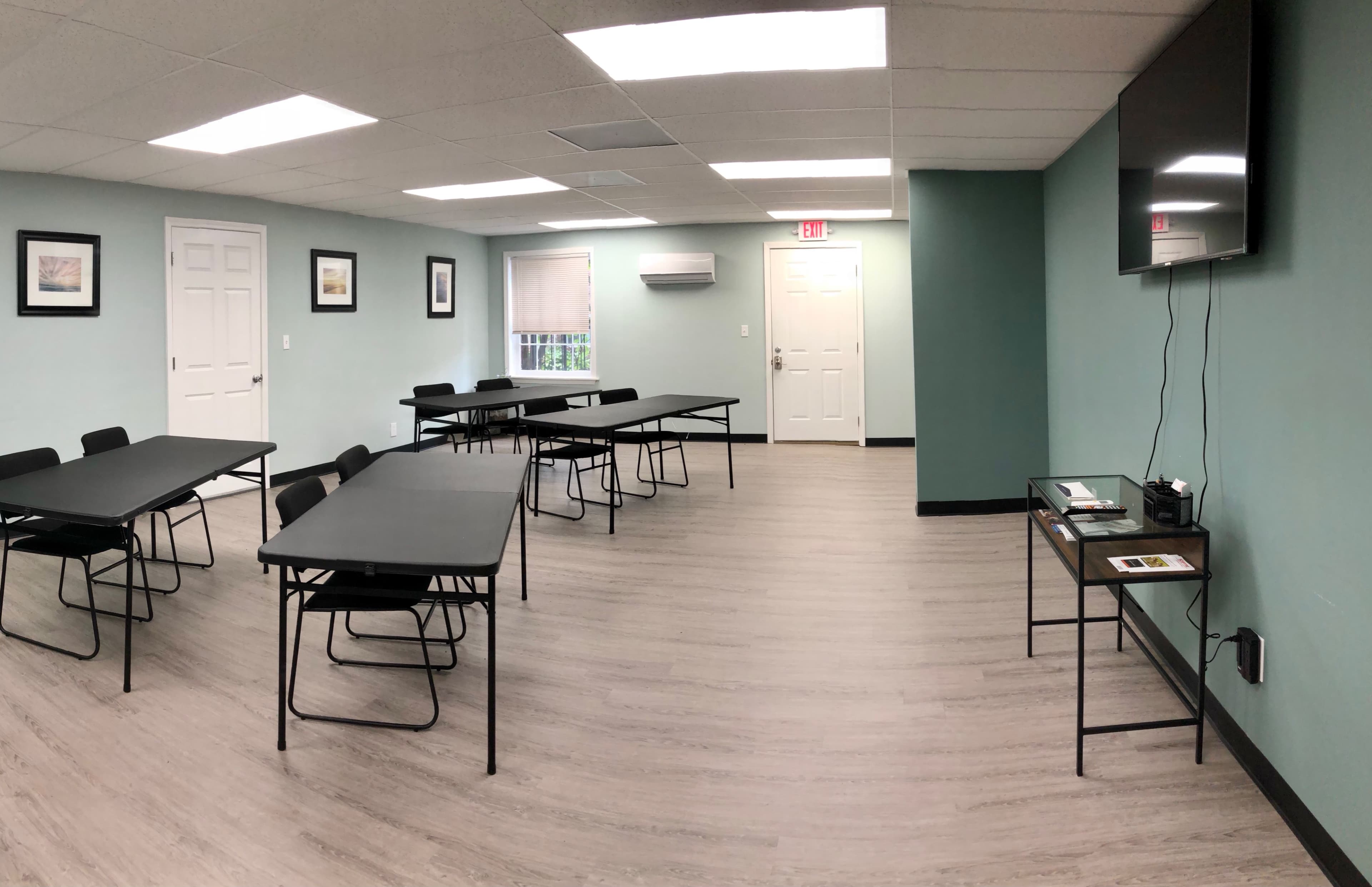 A meeting room with several tables and chairs arranged throughout, featuring light green walls and a wall-mounted television.
