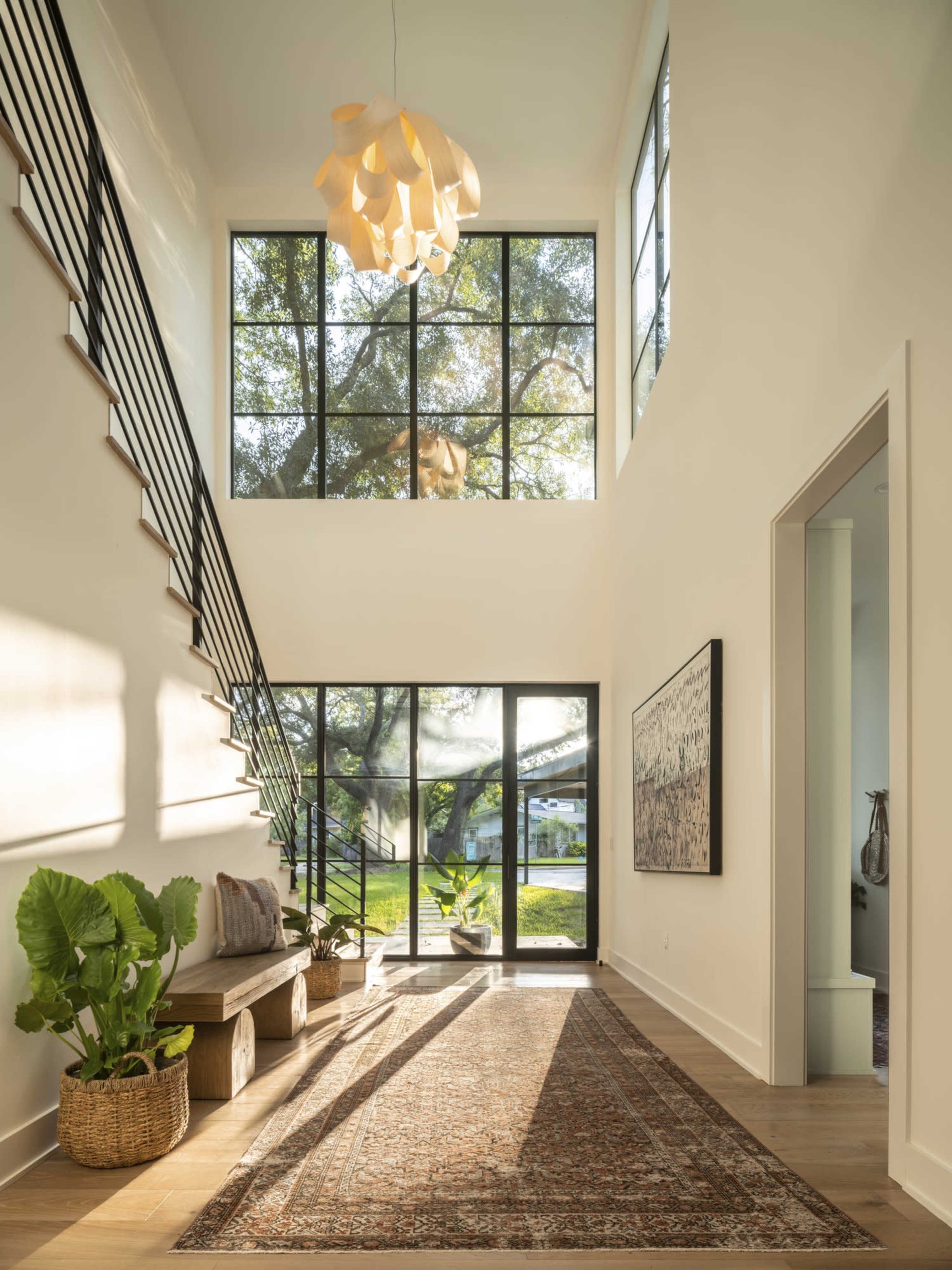 A bright entryway features a large staircase, tall windows, and wooden benches, complemented by a rug and potted plants.