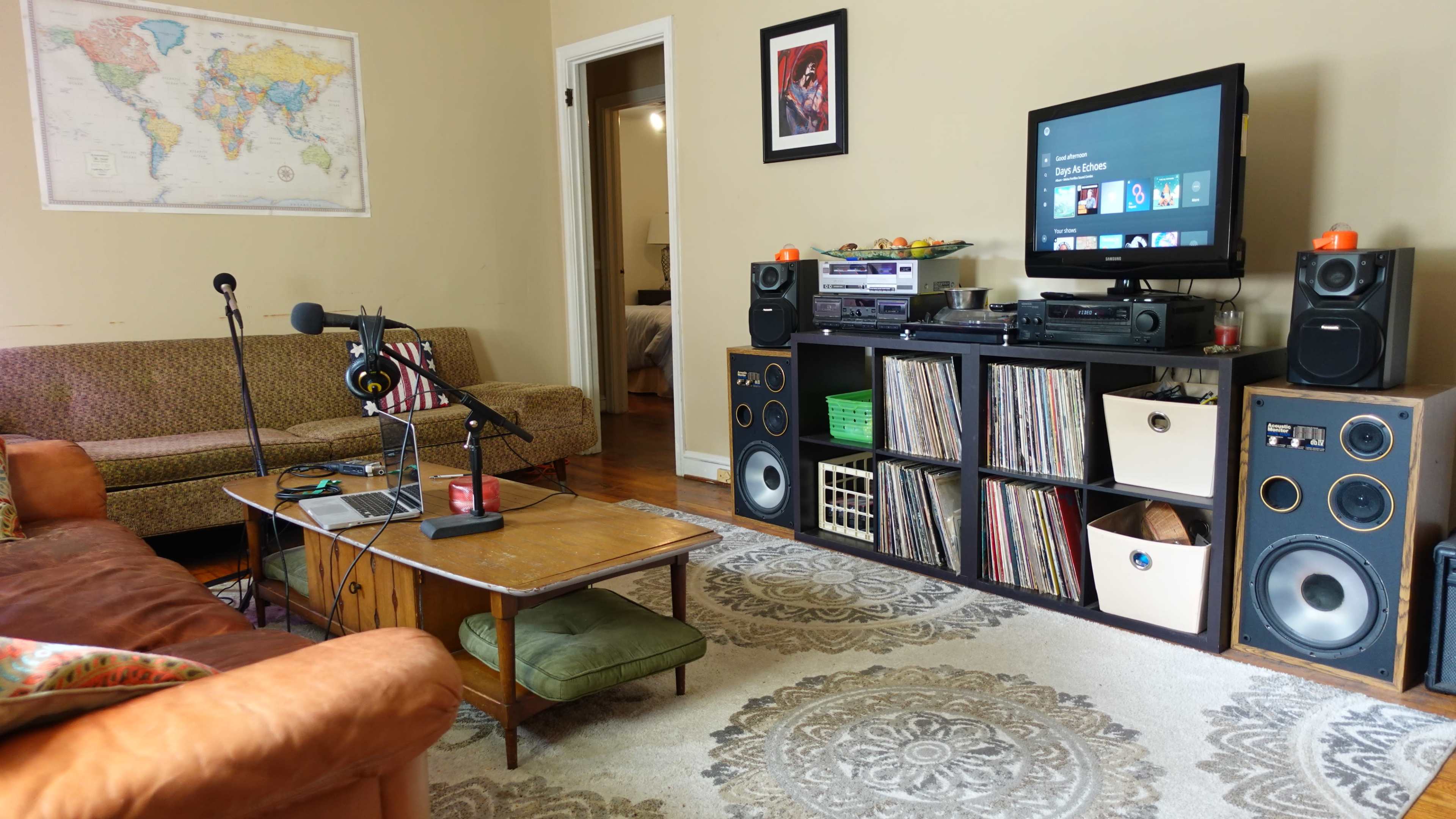 A living room features a muted color scheme, with a couch, a coffee table, a microphone on a stand, a television, and shelves holding vinyl records and audio equipment.