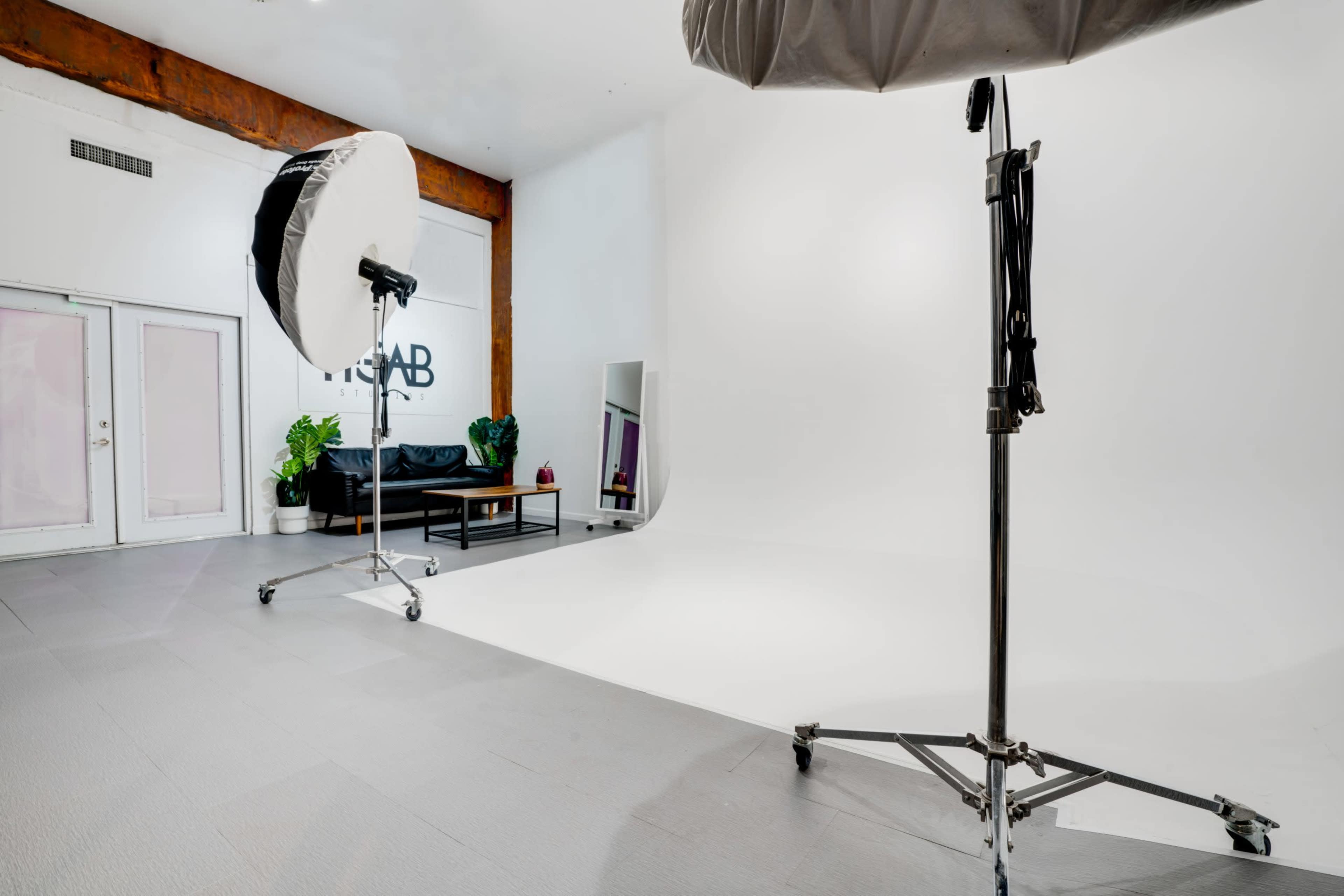 A spacious photography studio features a white backdrop, a black couch, and lighting equipment on rolling stands.
