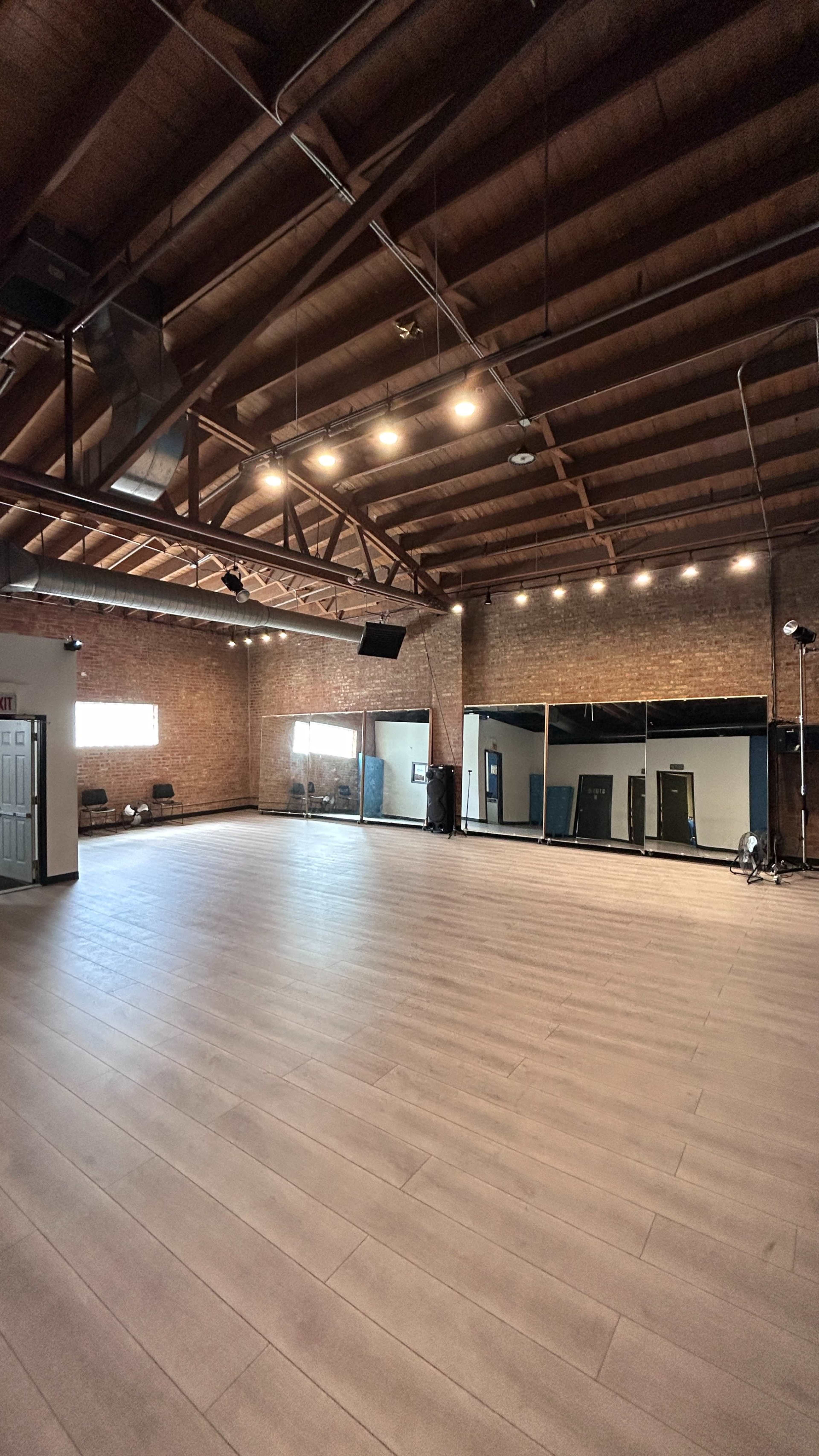 The image shows a spacious interior room with wooden flooring, exposed brick walls, and a high ceiling featuring beams and lights.