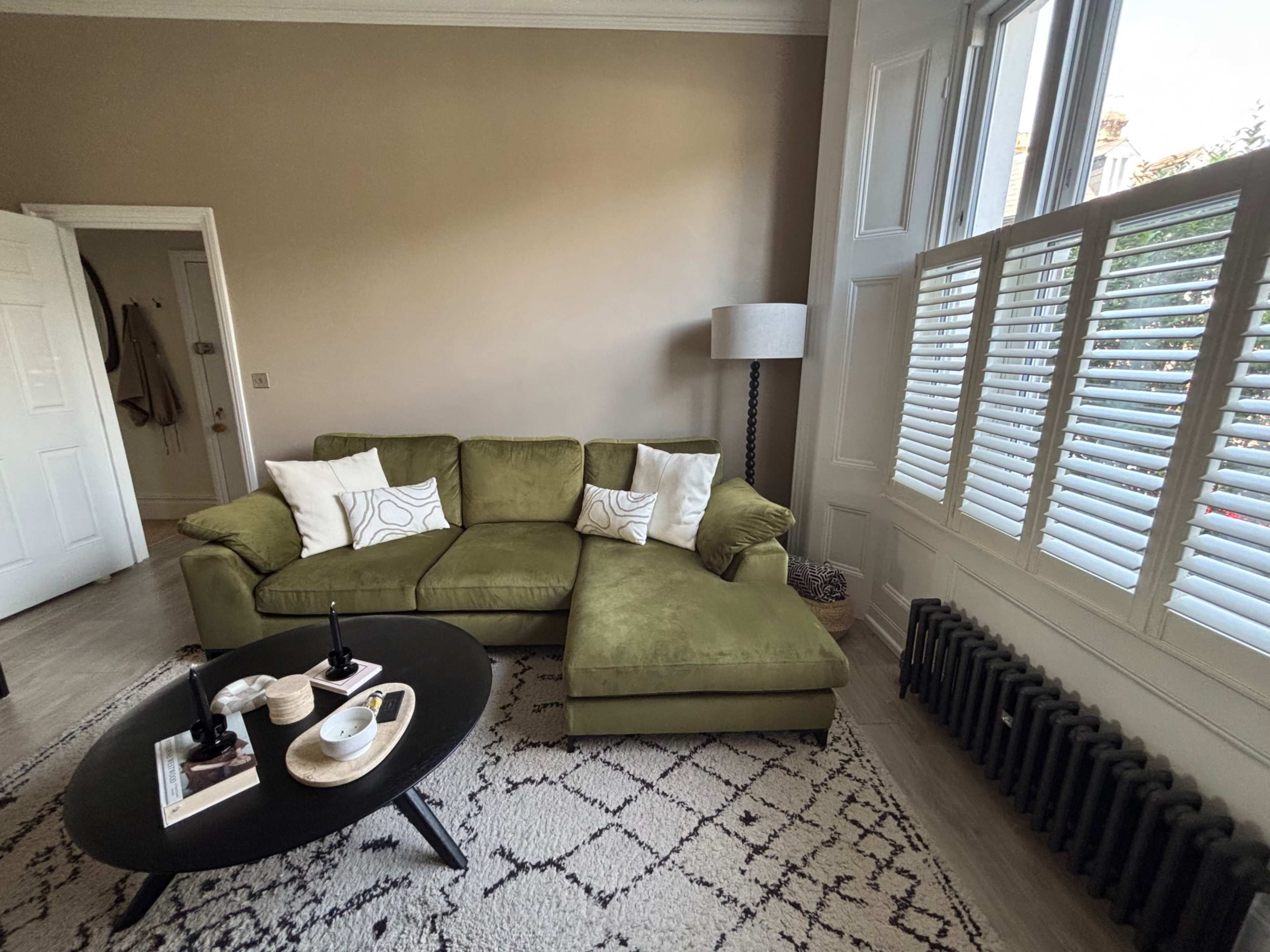 The image shows a living room with a green sectional sofa, two decorative pillows, a black coffee table, and a lamp beside a large window with shutters.