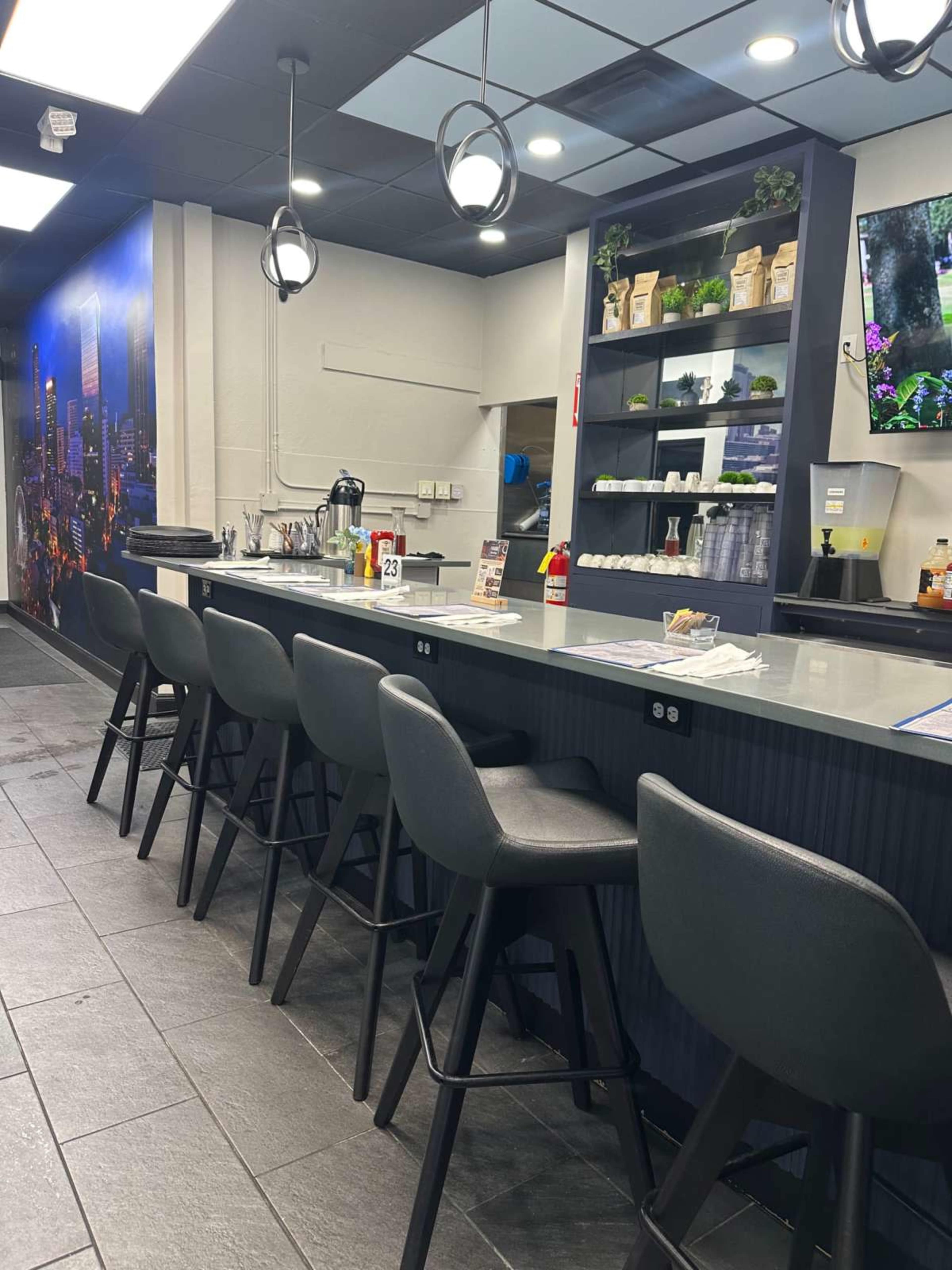 The image depicts a modern café interior with a long, dark counter, several high stools, and shelves displaying various decorative items, all set against a backdrop of a city skyline mural.