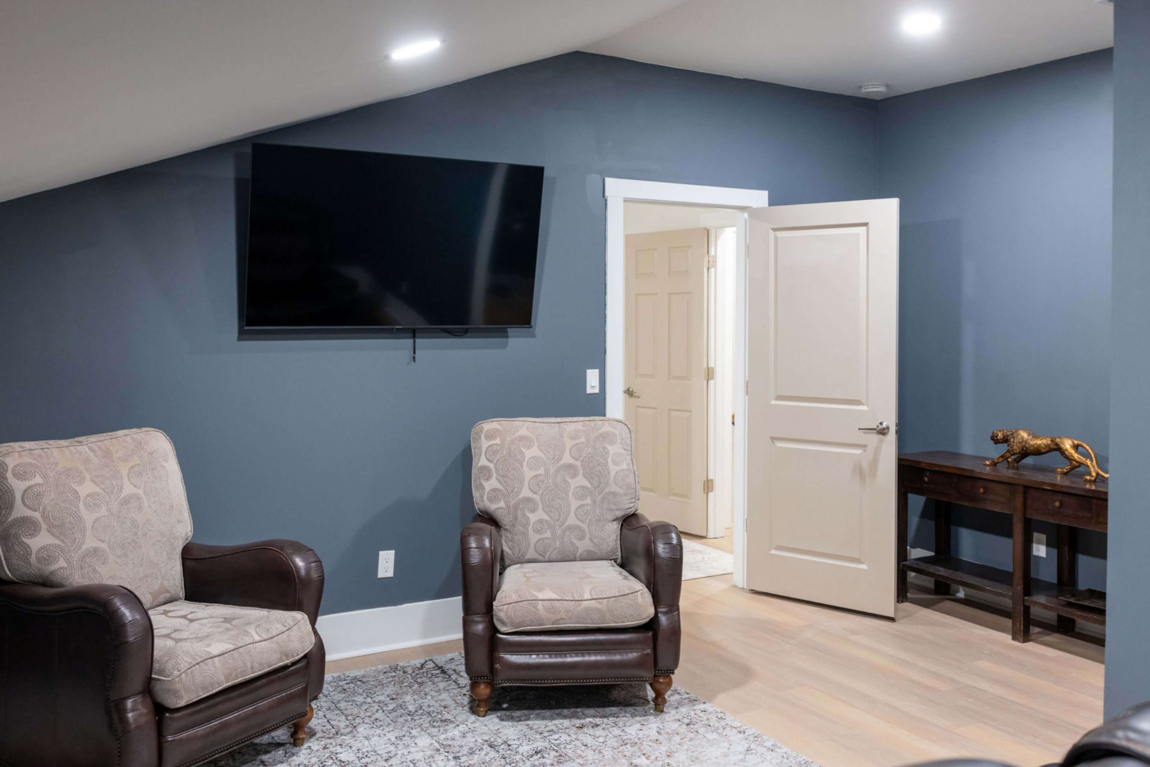 A cozy room features two armchairs facing a mounted television, with a side table and an open door leading to another space.