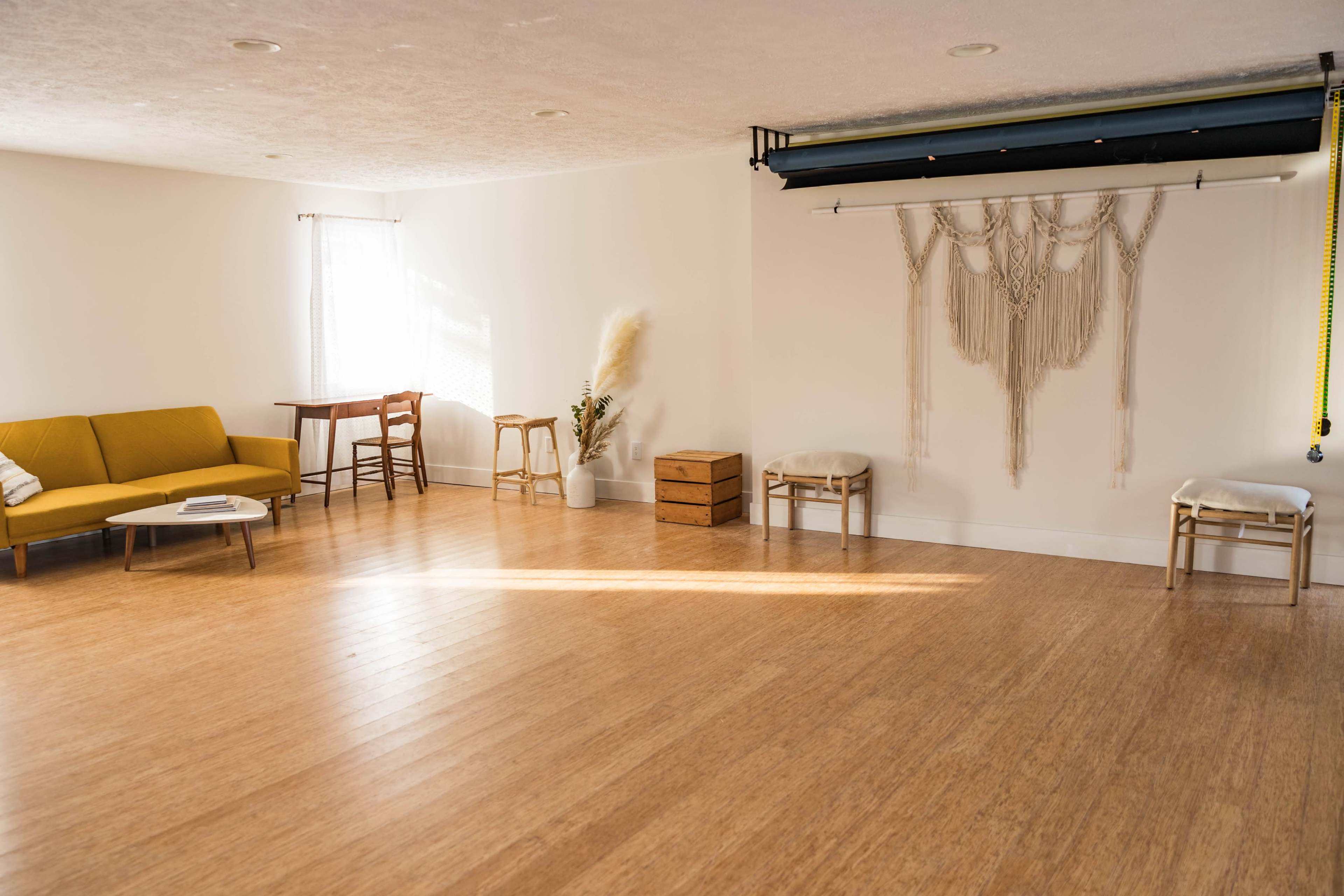 A minimalistic room with a yellow sofa, two stools, a wooden box, and wall decor made of macramé.