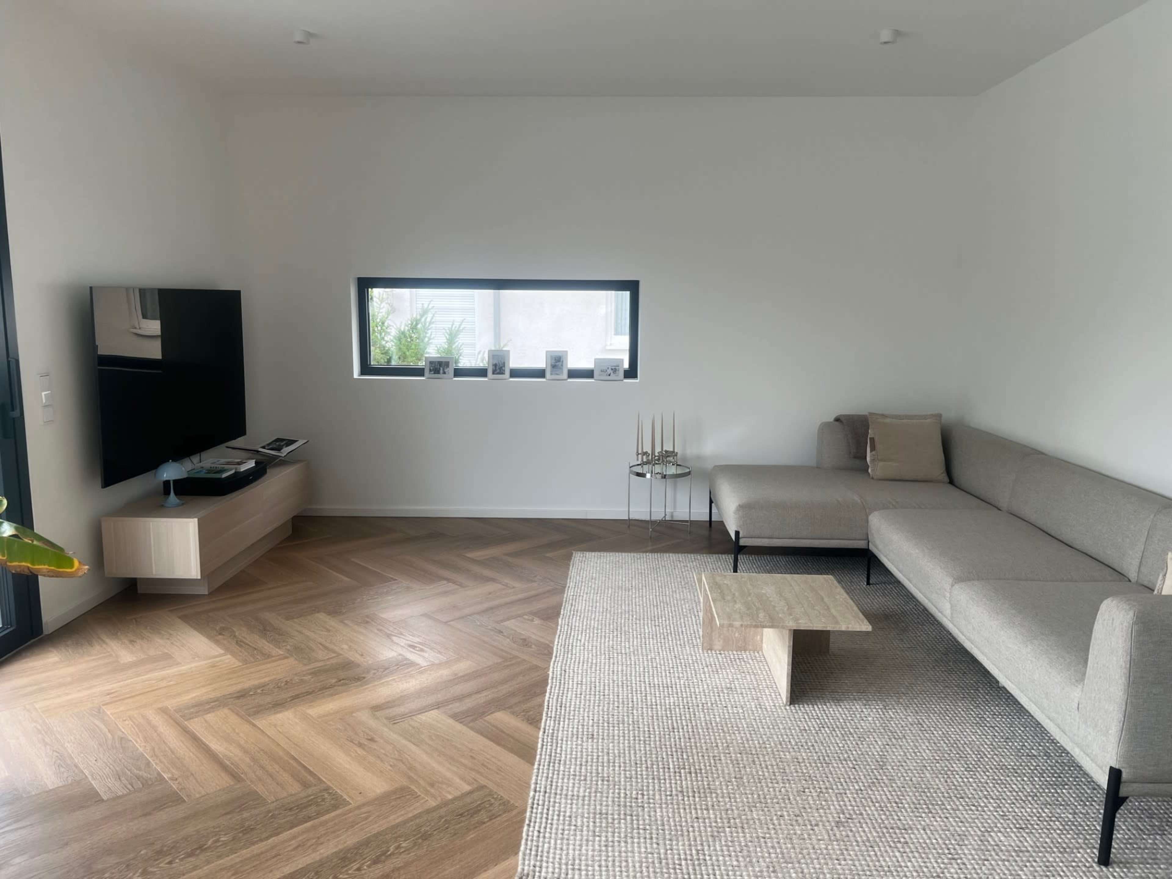 The image shows a modern living room featuring a light gray sofa, a wooden coffee table, a flat-screen TV on a side cabinet, and a small window with plants outside.