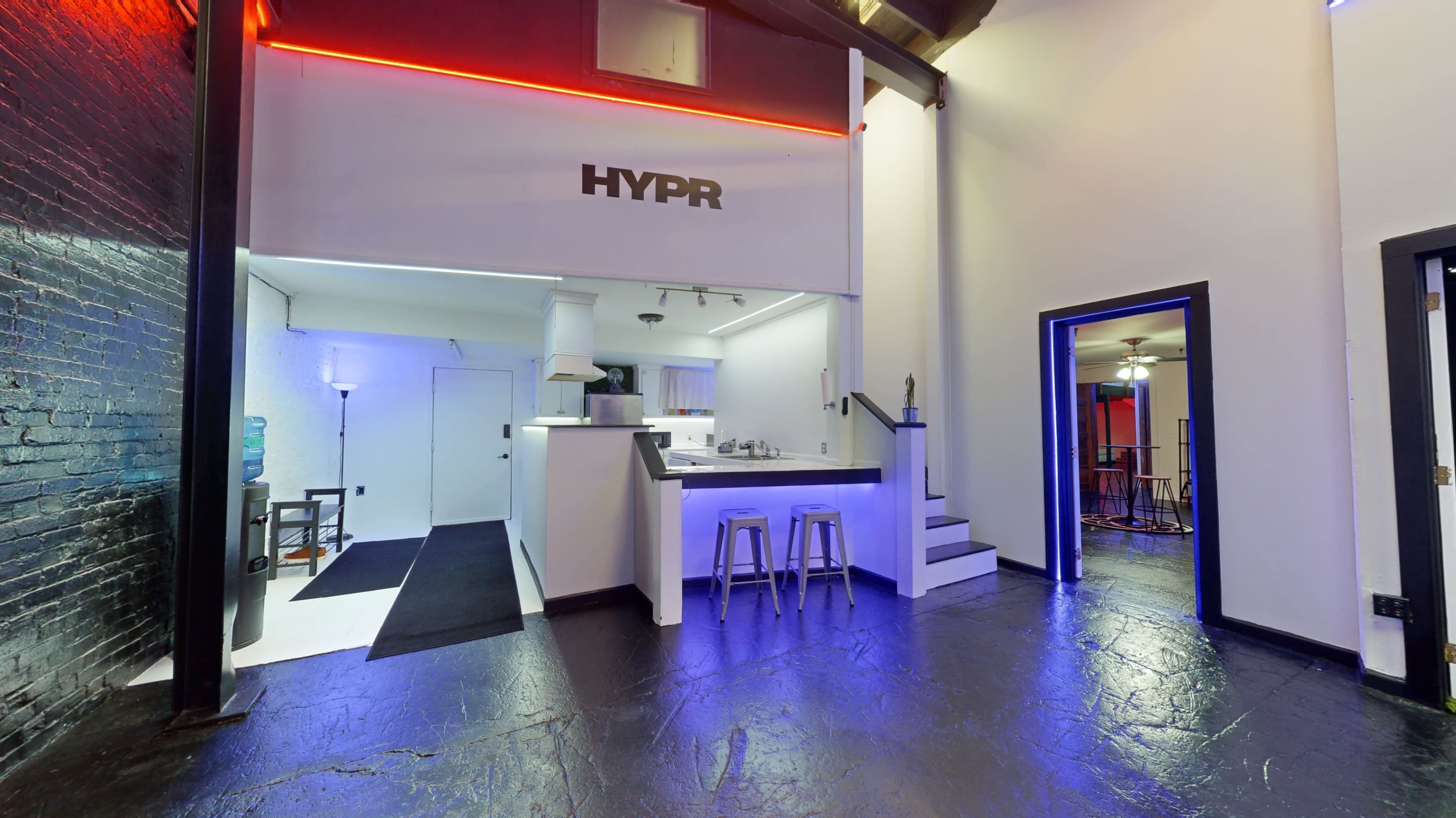 A modern, open interior space with a kitchen area, bar stools, and contrasting lighting effects on the walls.