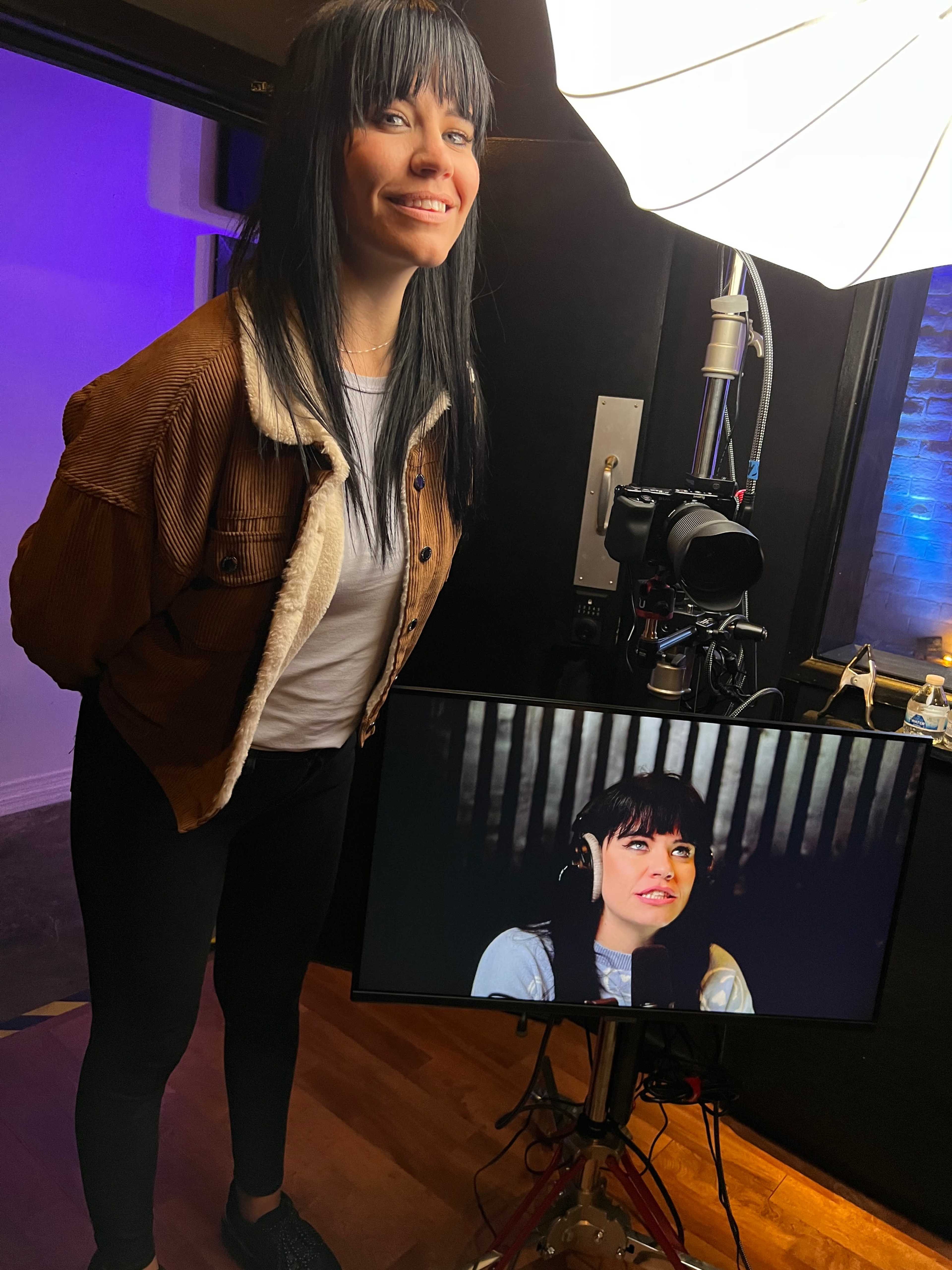 A woman stands beside a microphone and camera setup, looking at a monitor displaying her image.