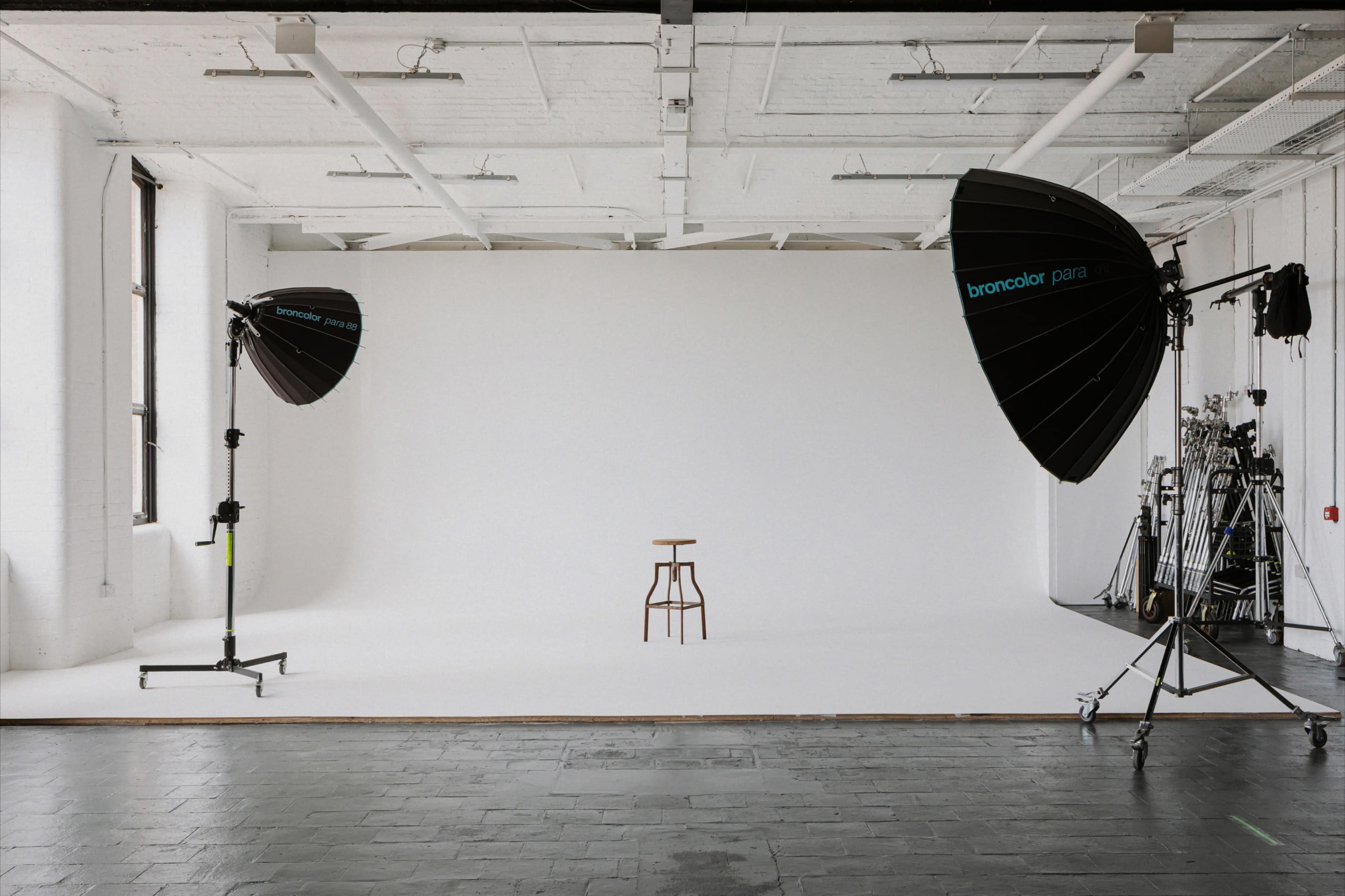A photography studio features a plain white backdrop with a single wooden stool centered in the foreground, flanked by large lighting setups.