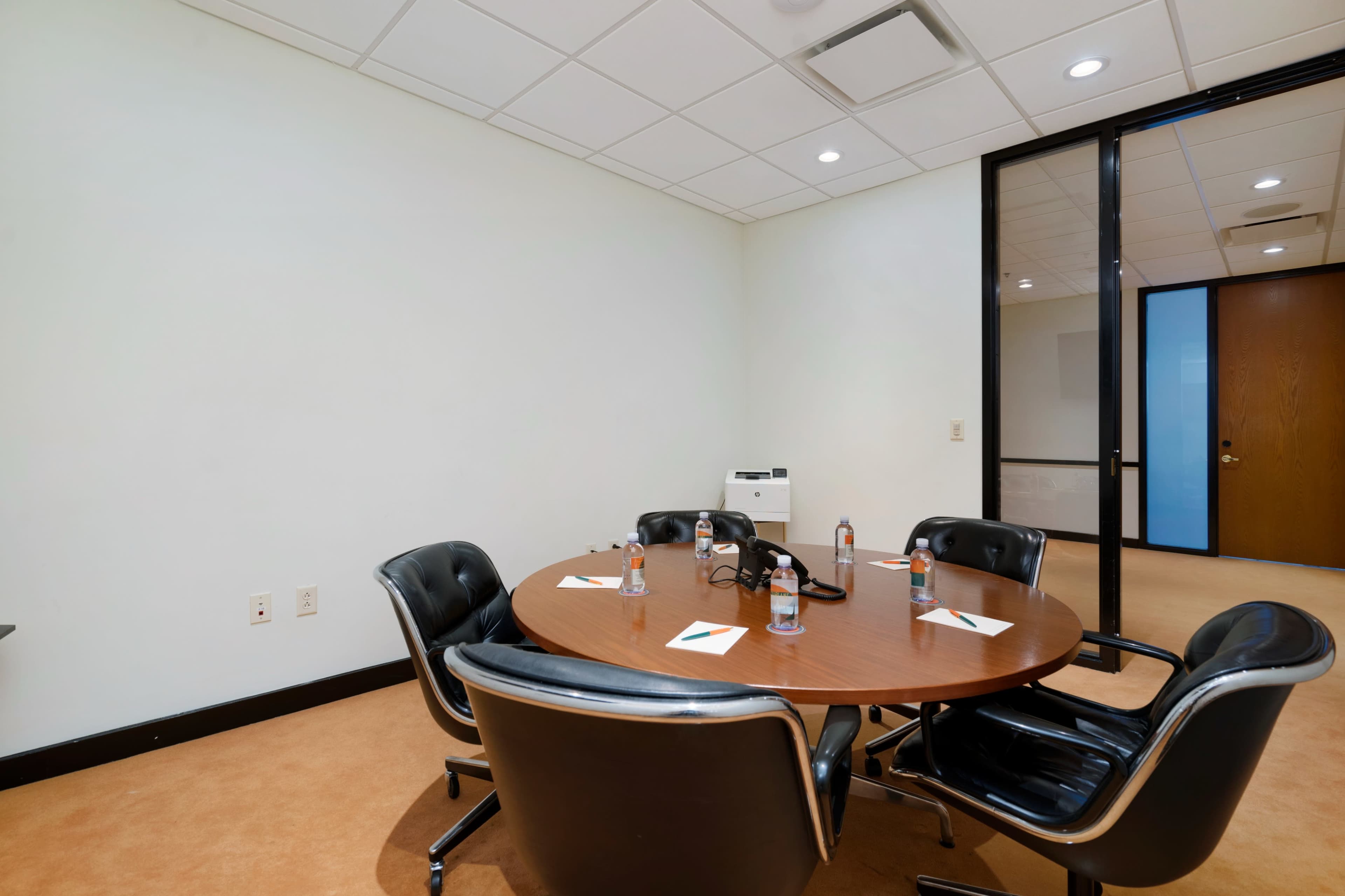 A round wooden conference table is set with four black chairs, water bottles, and notepads in a simple meeting room.