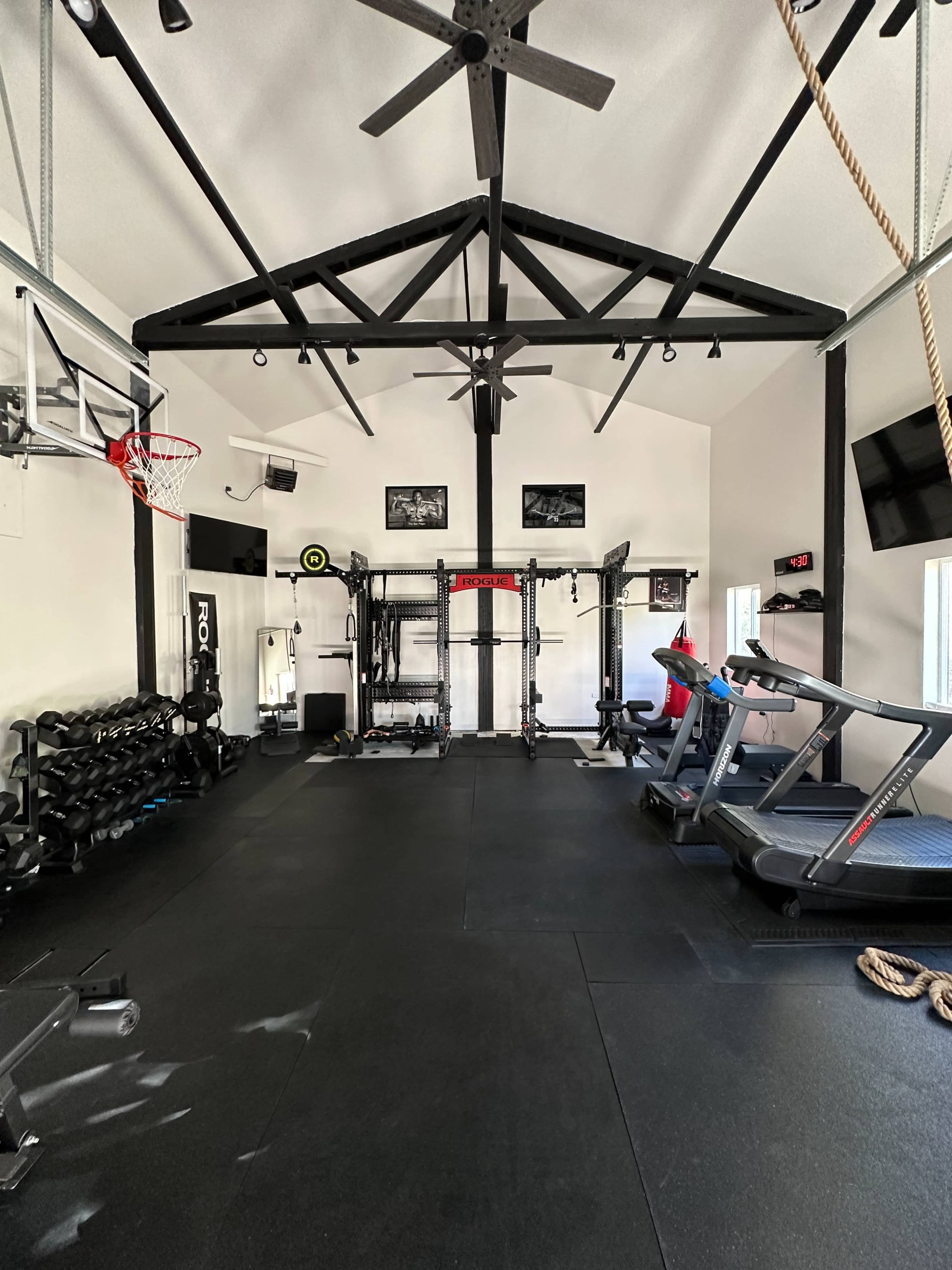 The image shows an interior of a home gym featuring various exercise equipment, including dumbbells, a treadmill, and a weight rack, with a basketball hoop mounted on the wall.