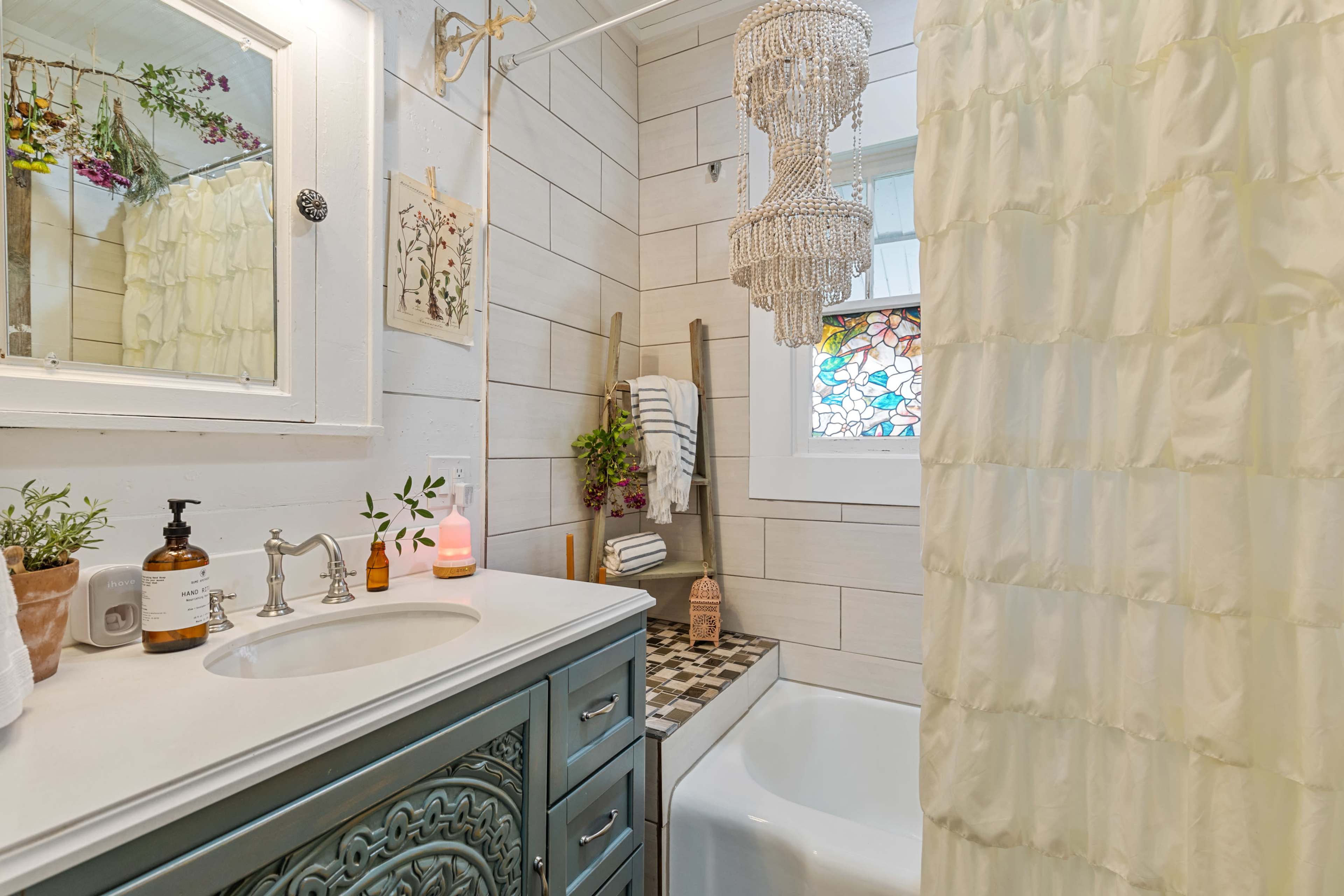 The bathroom features a clawfoot tub, a decorative vanity with a blue finish, and a chandelier above, complemented by a stained glass window and decorative plants.