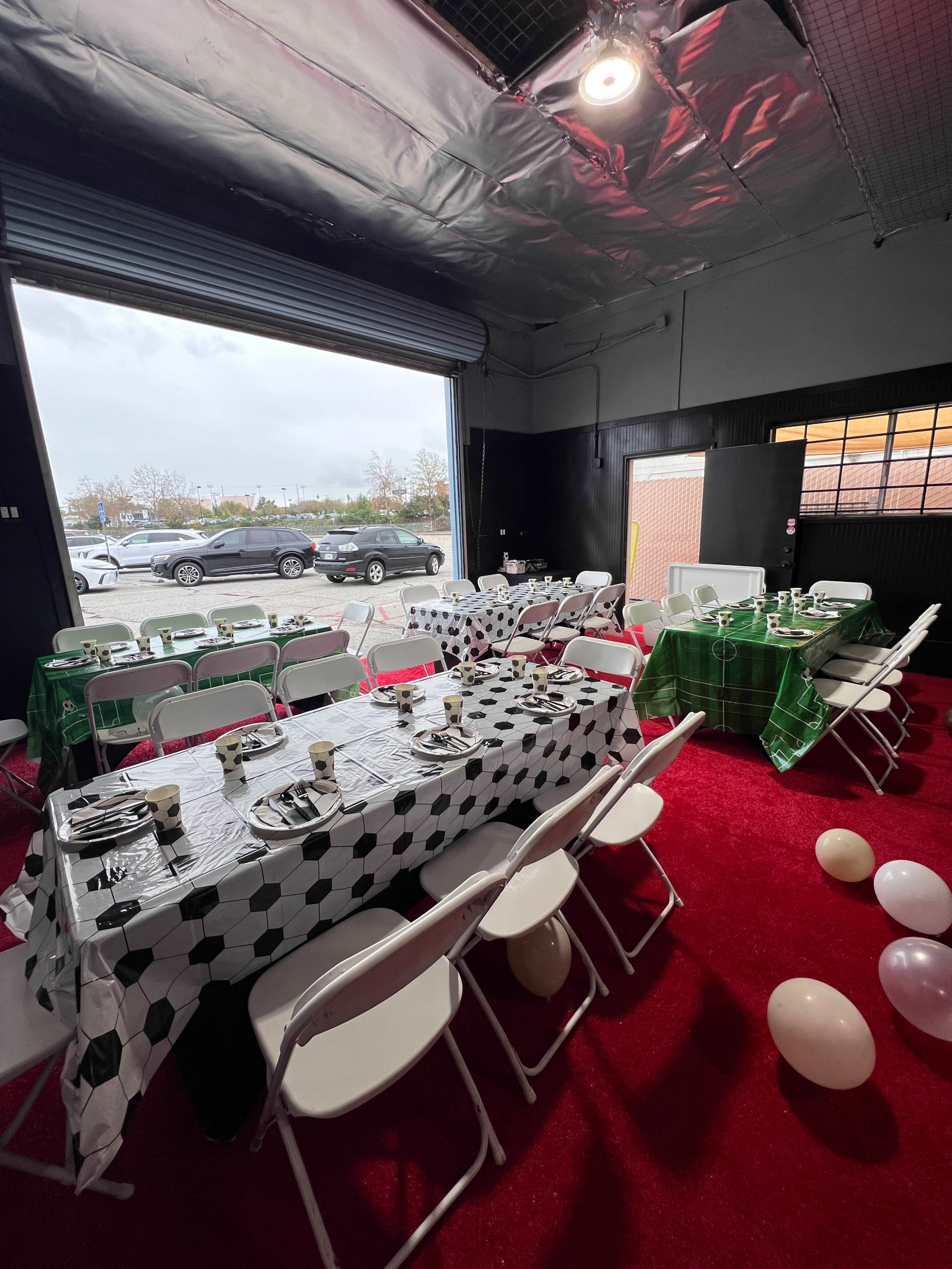 The image shows a decorated room with tables set for a gathering, featuring soccer-themed tablecloths and chairs, and a view of parked cars through an open door.