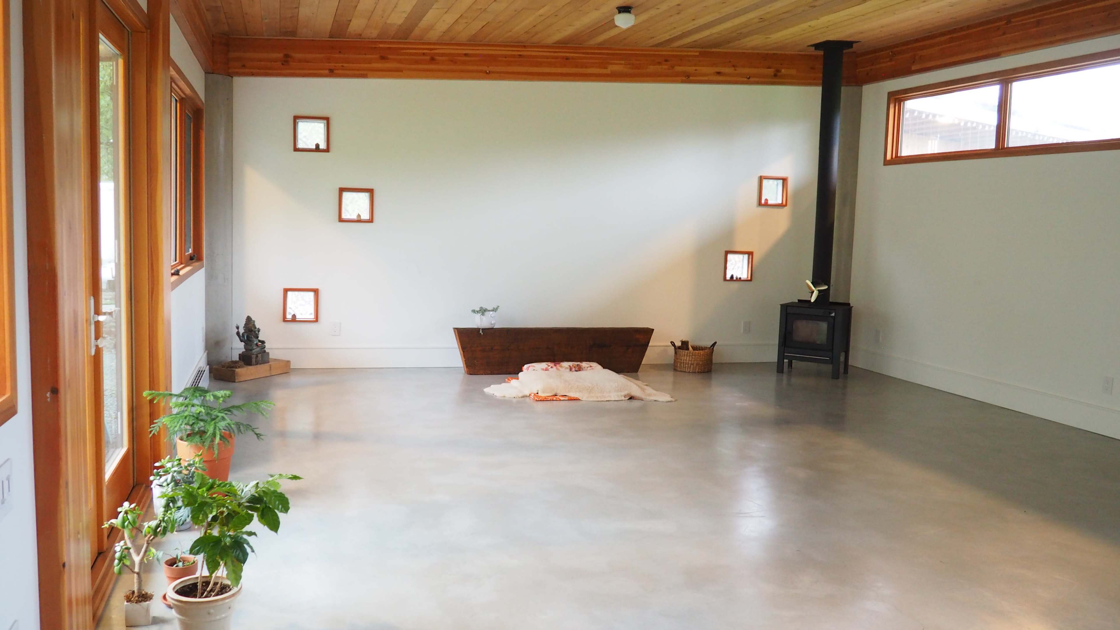 The room features a polished concrete floor, wooden ceiling, a small stove, and several windows that frame minimalistic decor.