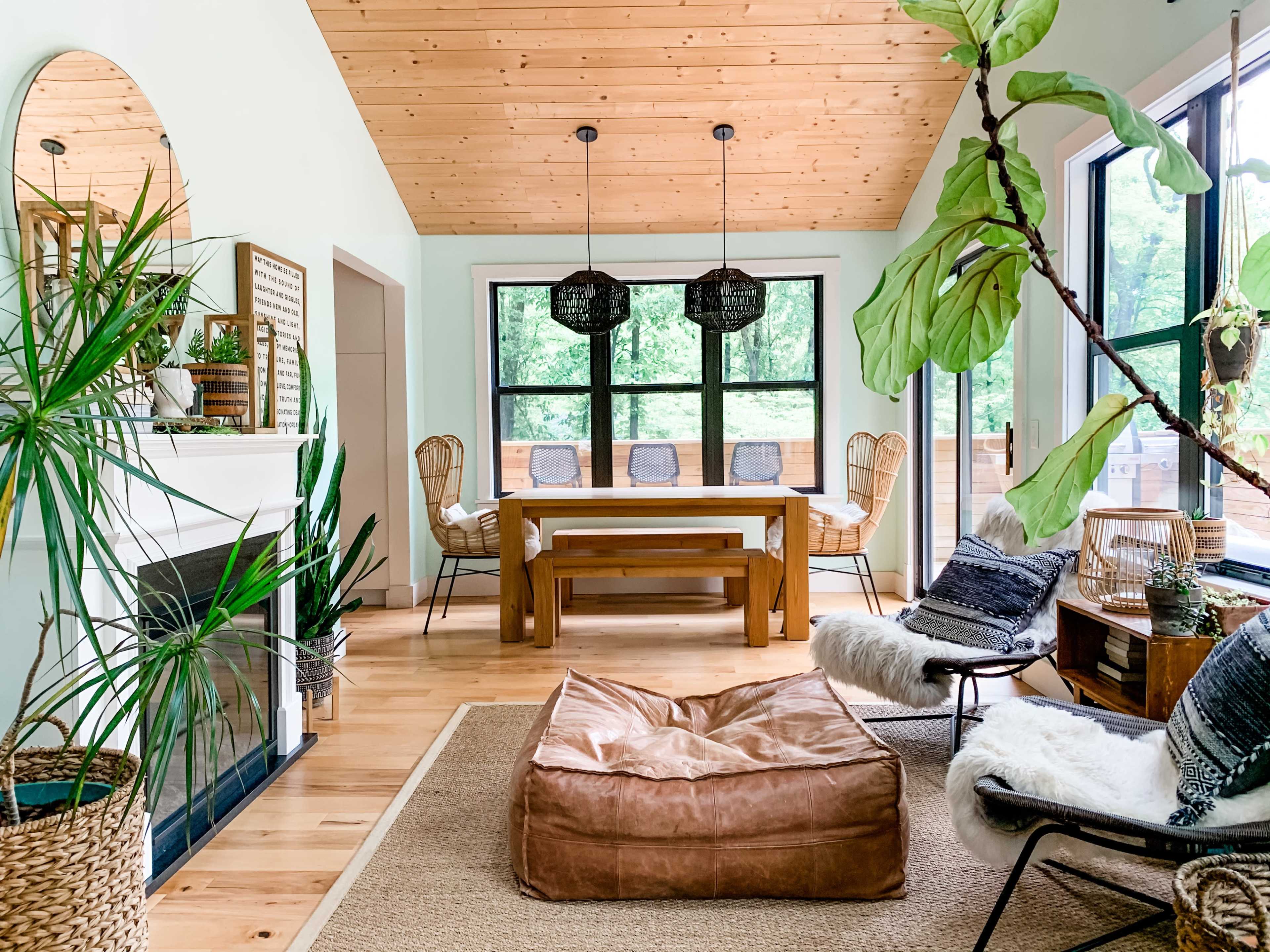 The image shows a modern dining area with a wooden table, rattan chairs, a large brown bean bag, and a fireplace, surrounded by greenery and large windows.