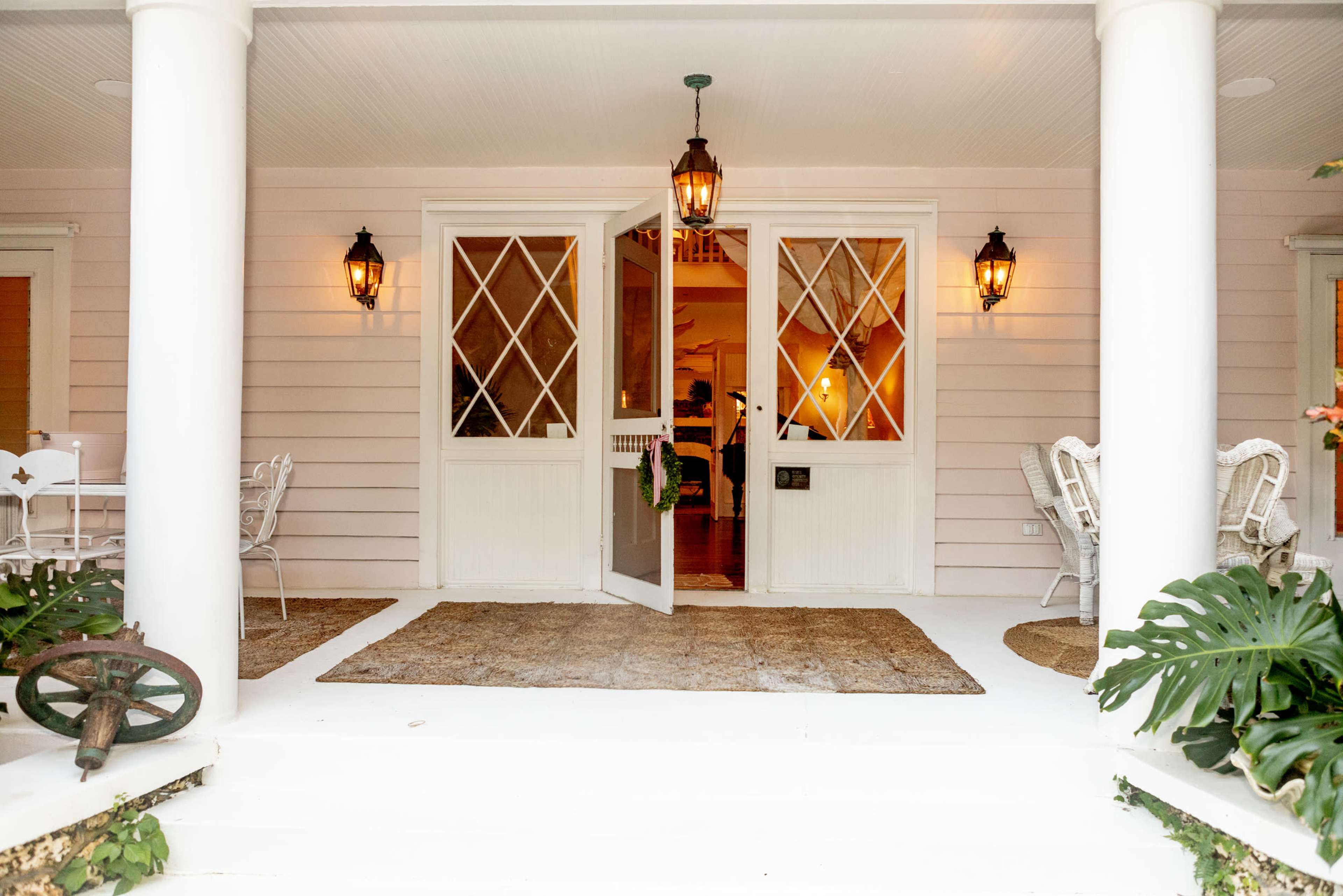The image shows a welcoming entryway with open double doors, two lanterns flanking the entrance, and outdoor seating arranged on a porch.