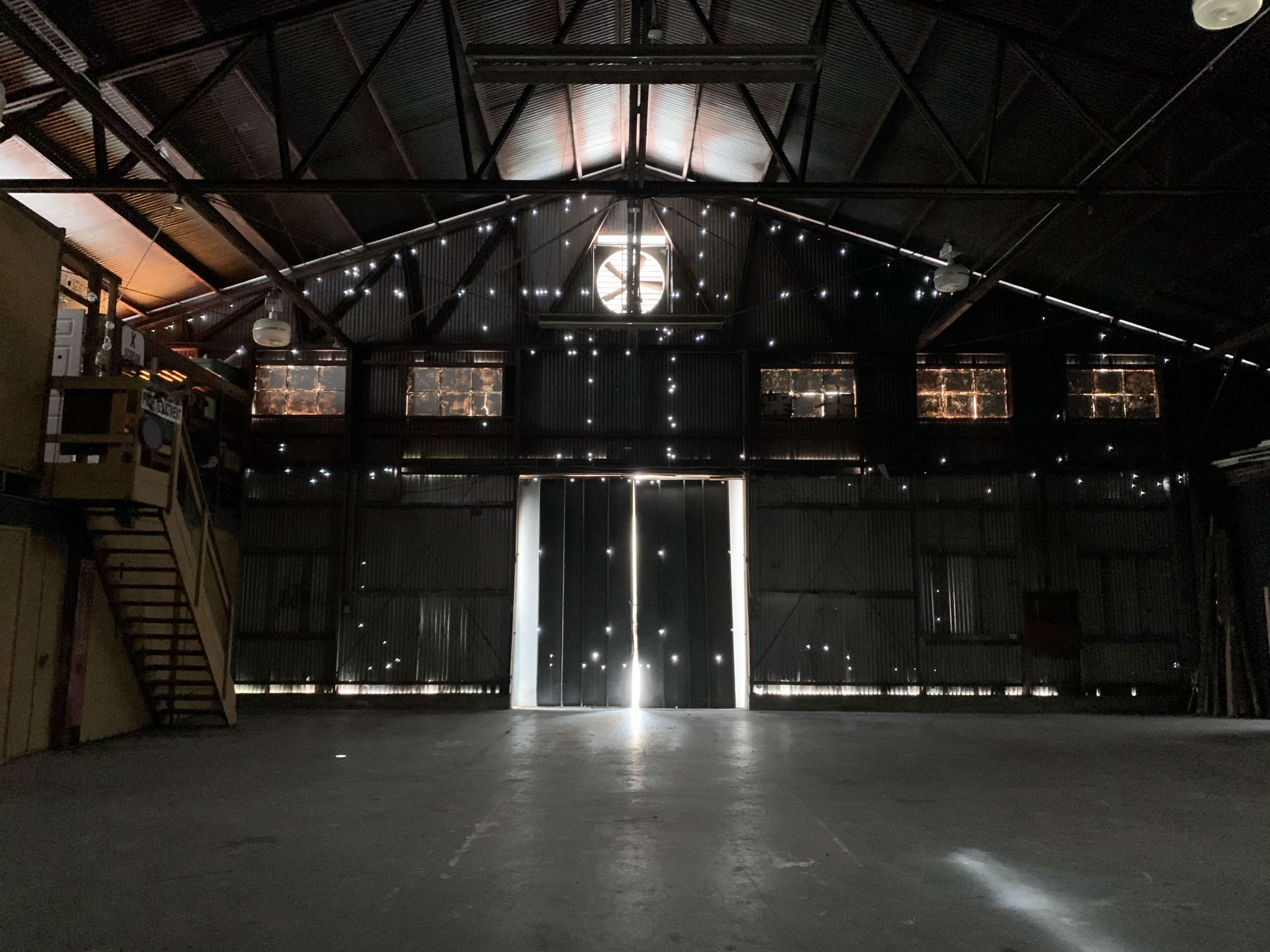 An empty warehouse features large doors at the back, with light streaming in through gaps in the walls and ceiling.