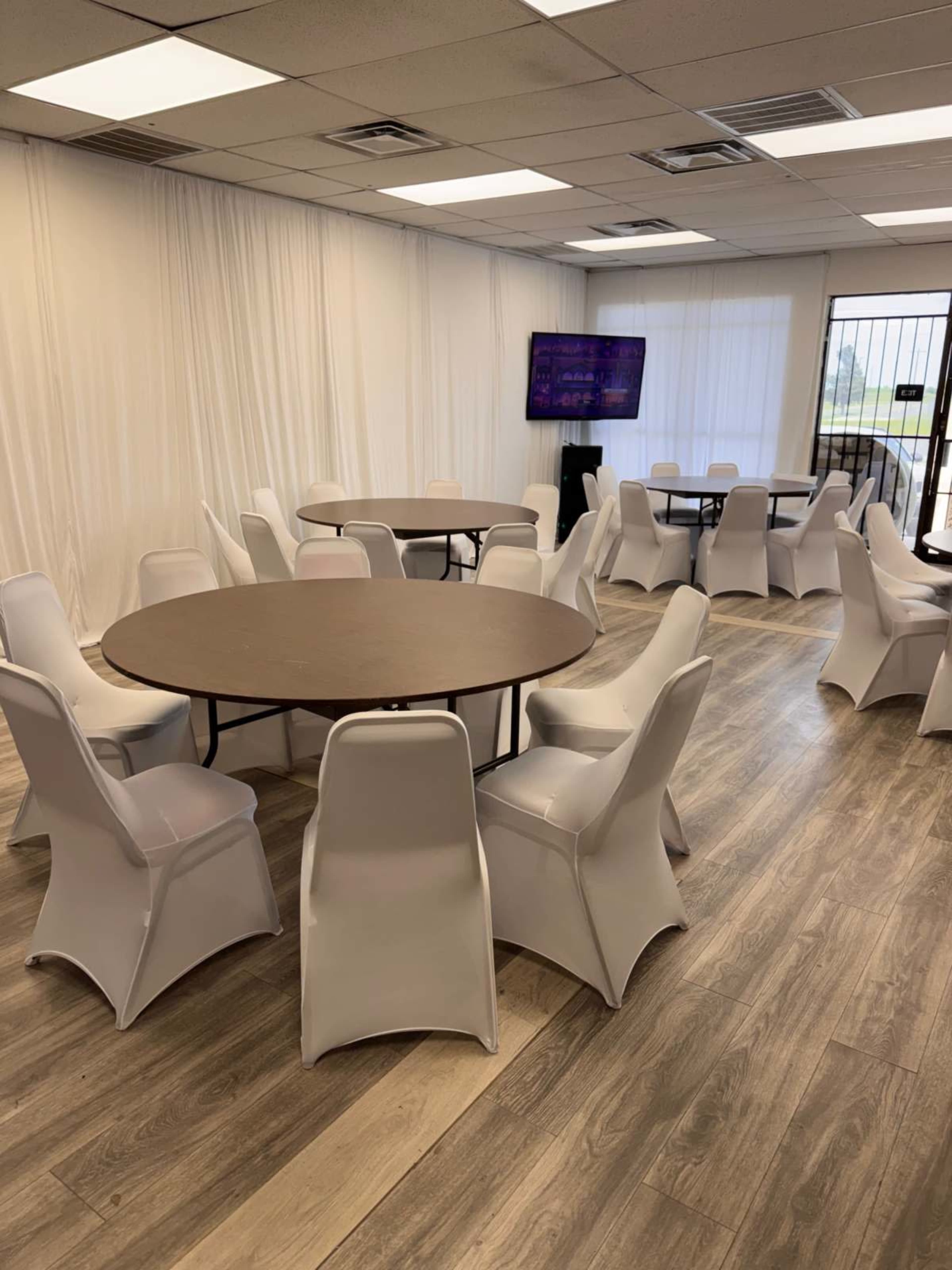 The image shows a spacious room with multiple round tables surrounded by white chairs, a television screen, and a light-colored backdrop.