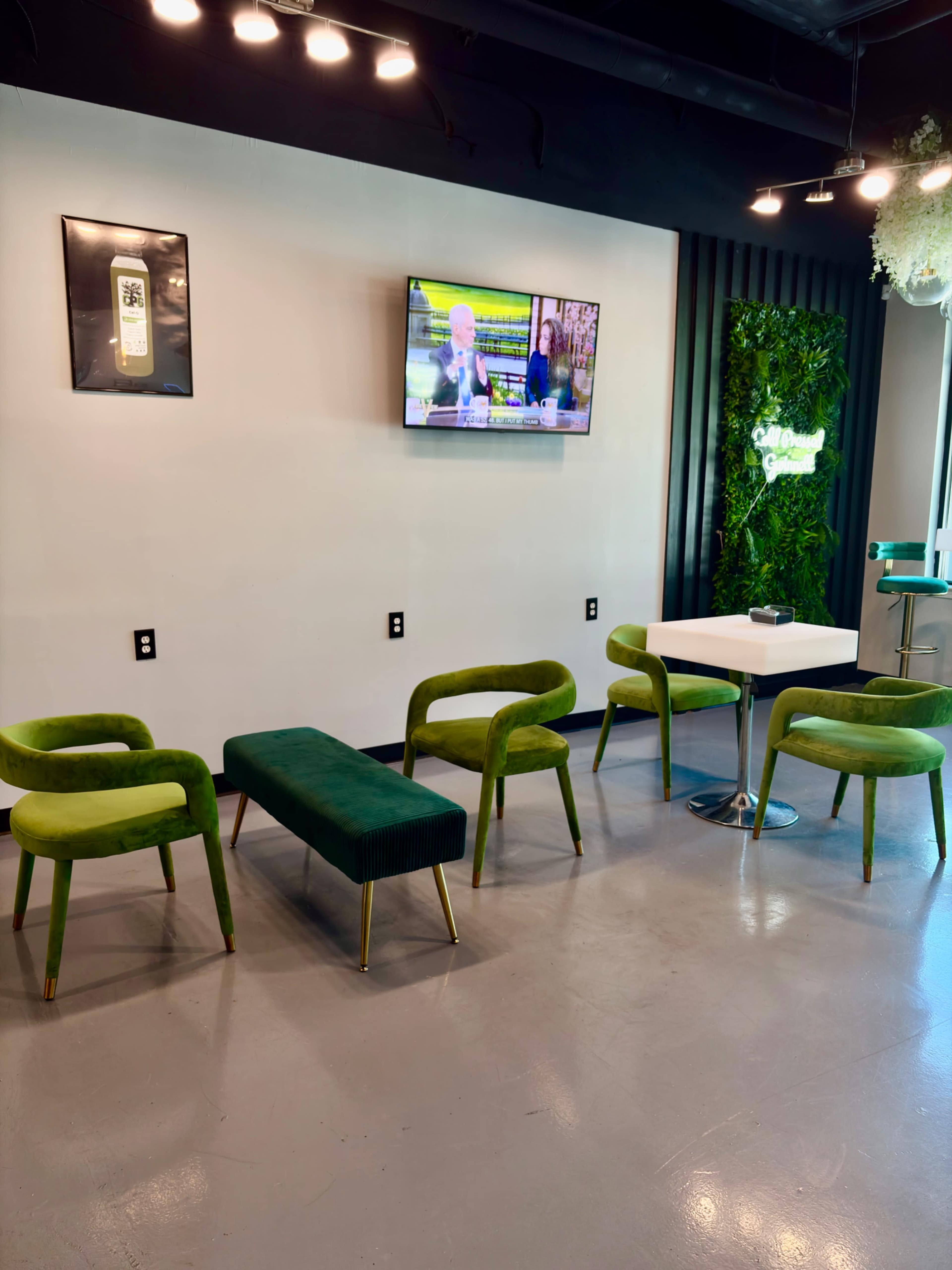 The image shows a modern waiting area featuring green upholstered chairs, a bench, a table, a television mounted on the wall, and a plant wall decoration.