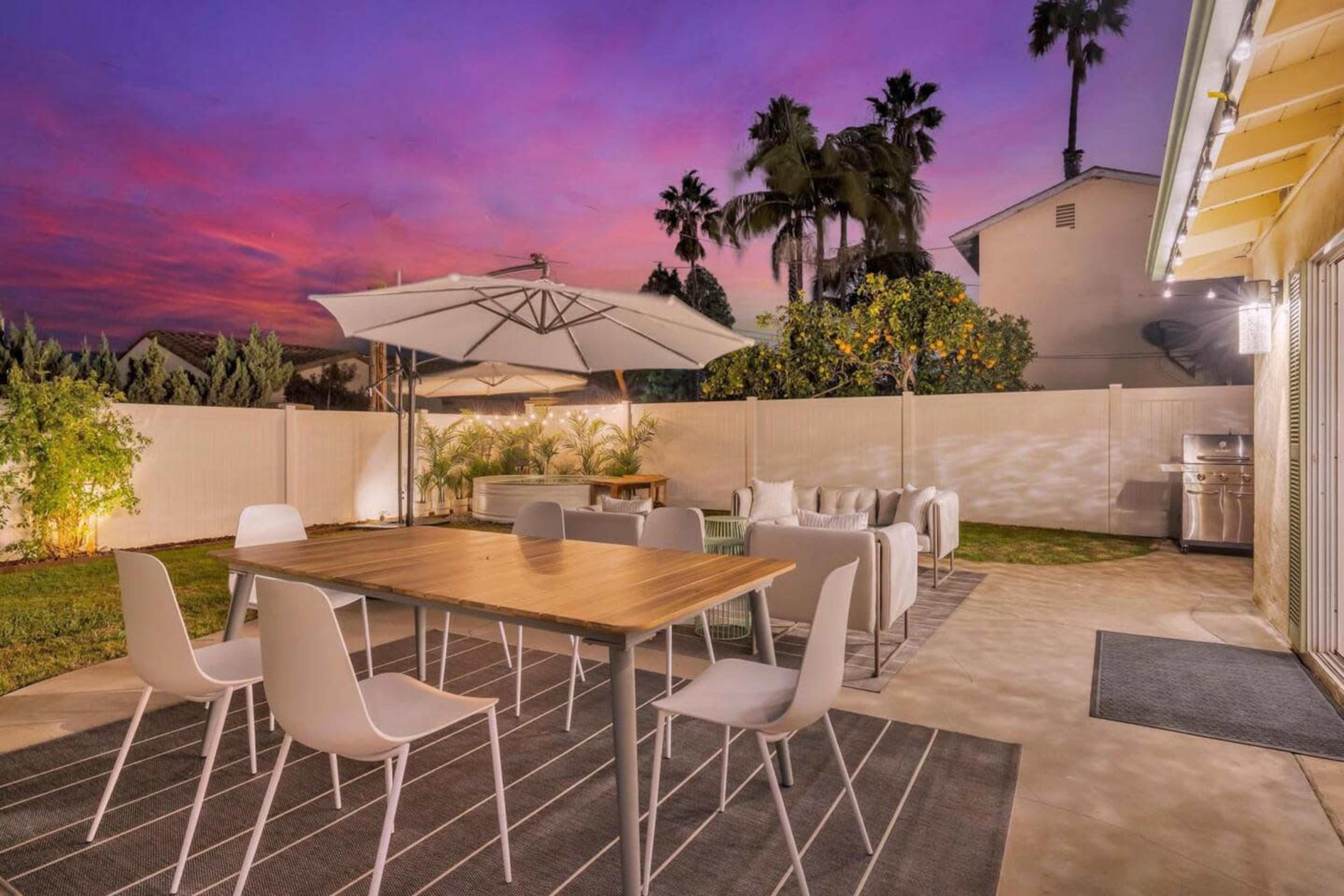 A backyard with a wooden dining table surrounded by white chairs, an umbrella, and a barbecue grill, set against a colorful sunset sky.