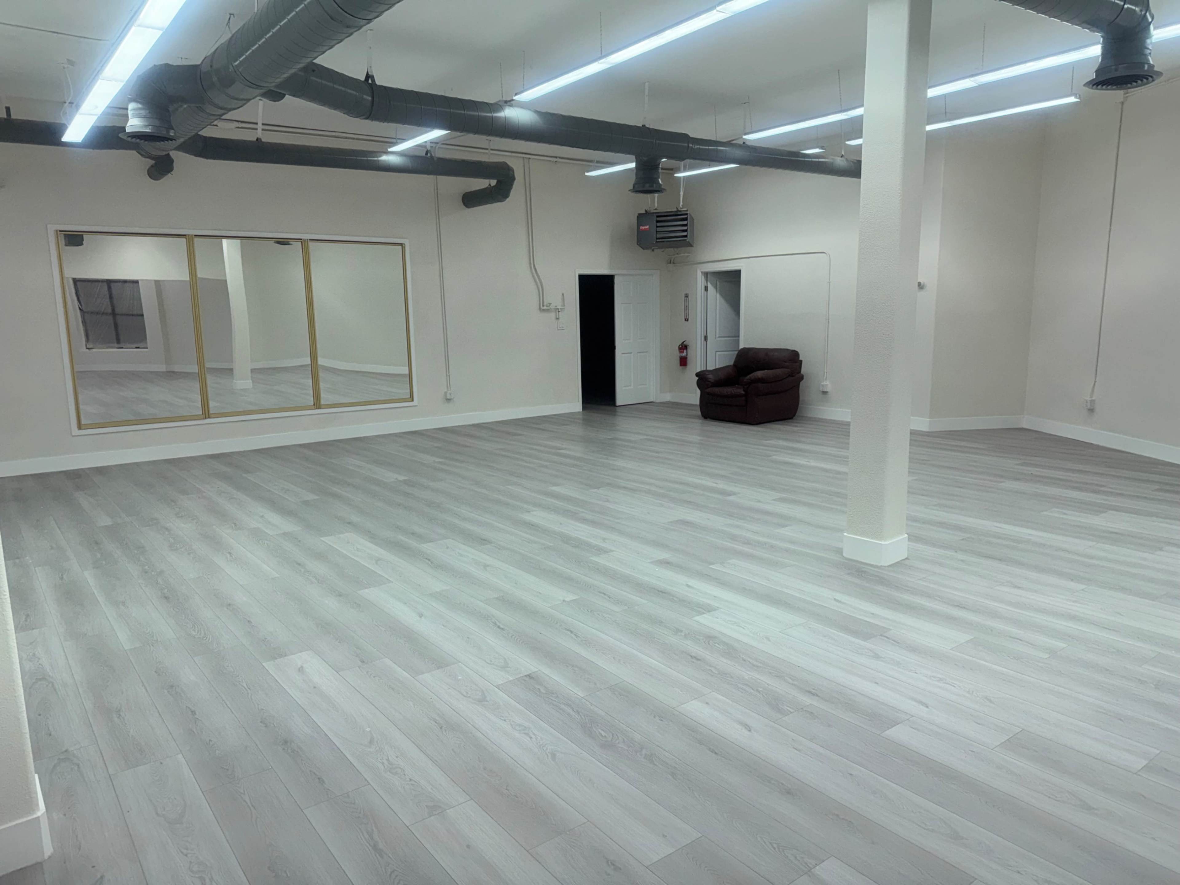 The image shows a spacious, empty room with light-colored flooring and a large mirror on one wall, featuring a single brown chair in the corner.