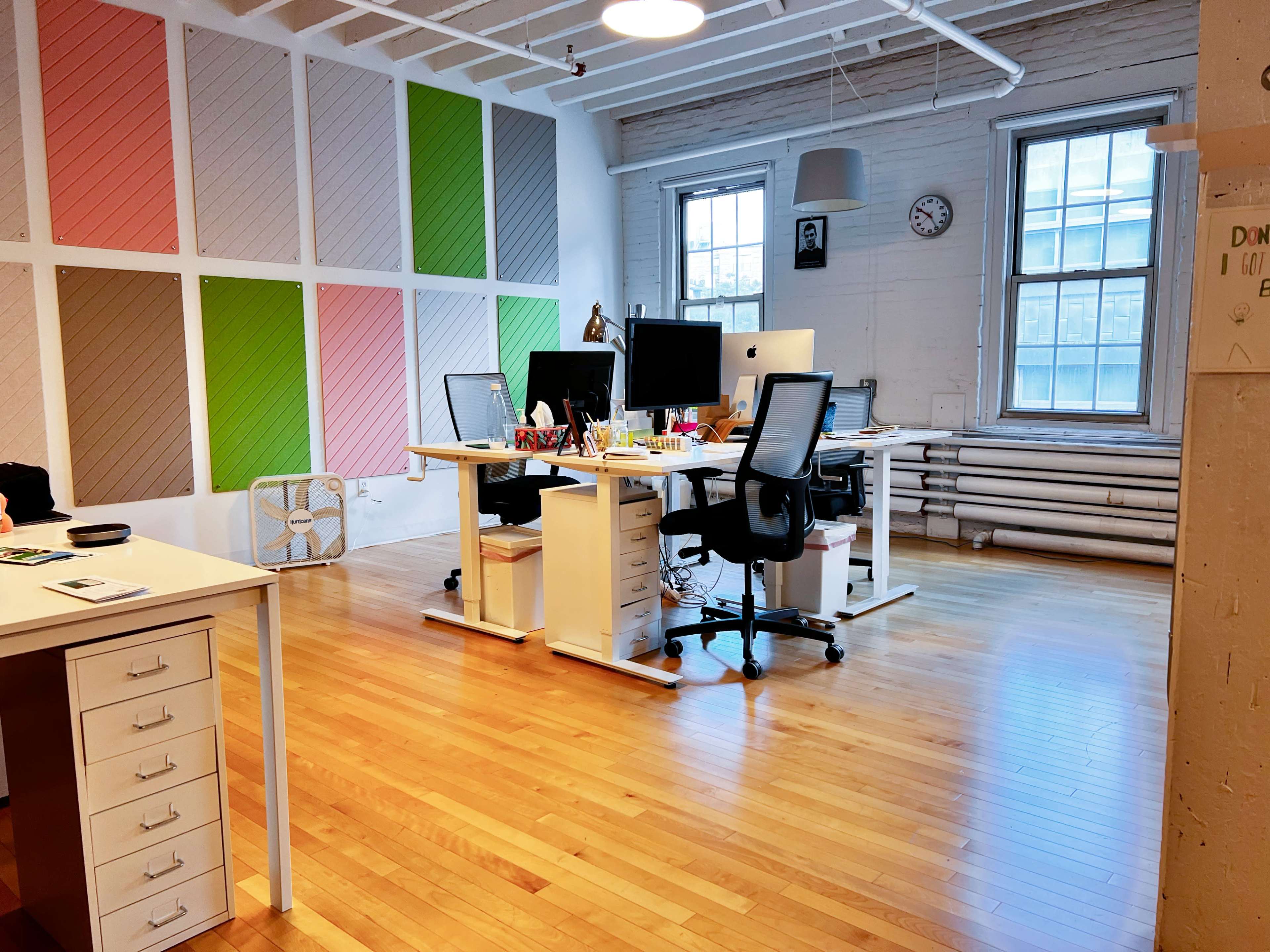 An office space with two desks featuring computers, surrounded by a wall with colored panels and large windows.