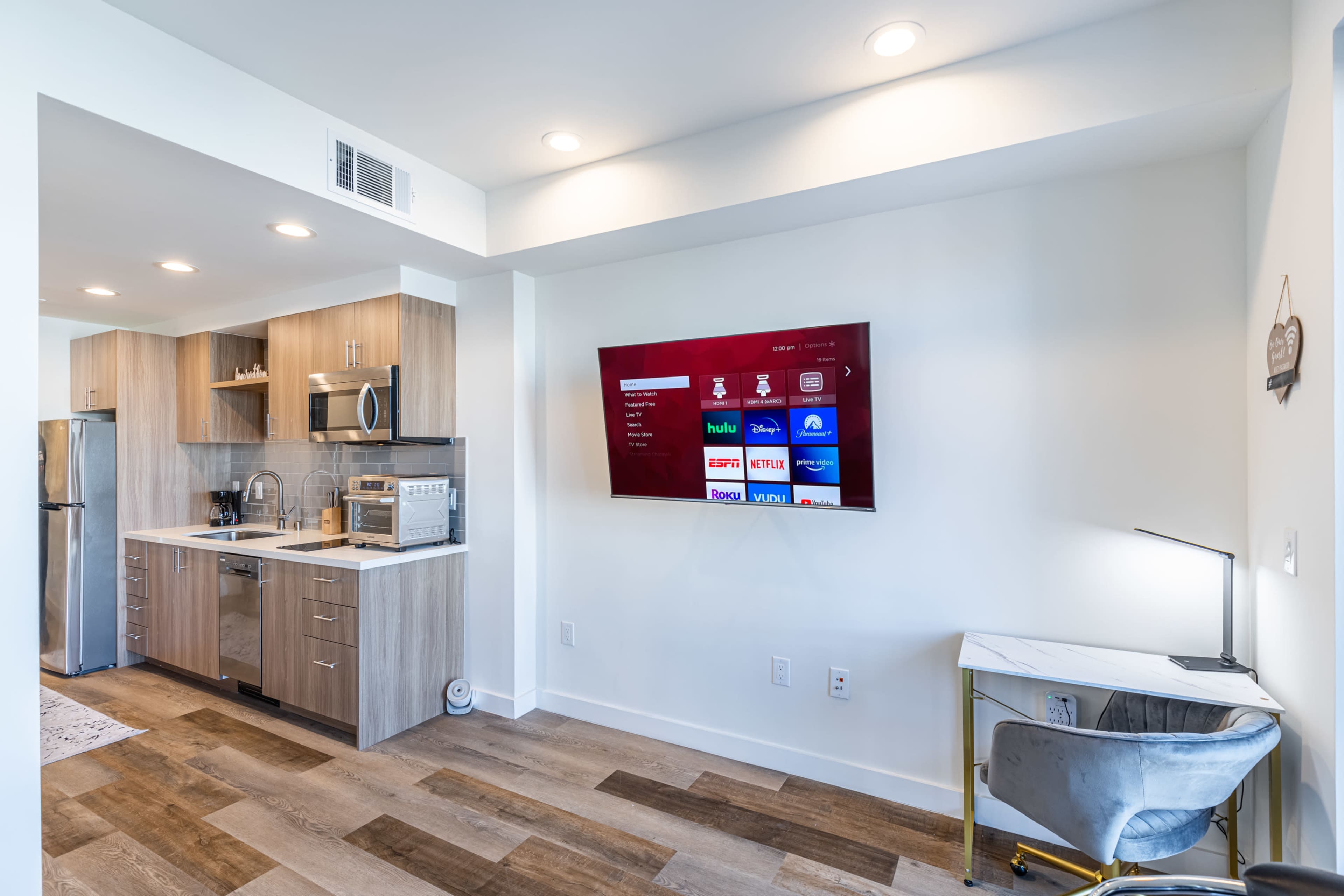 The image shows a modern kitchen and living area with a wall-mounted TV, a small desk with a chair, and contemporary cabinetry.