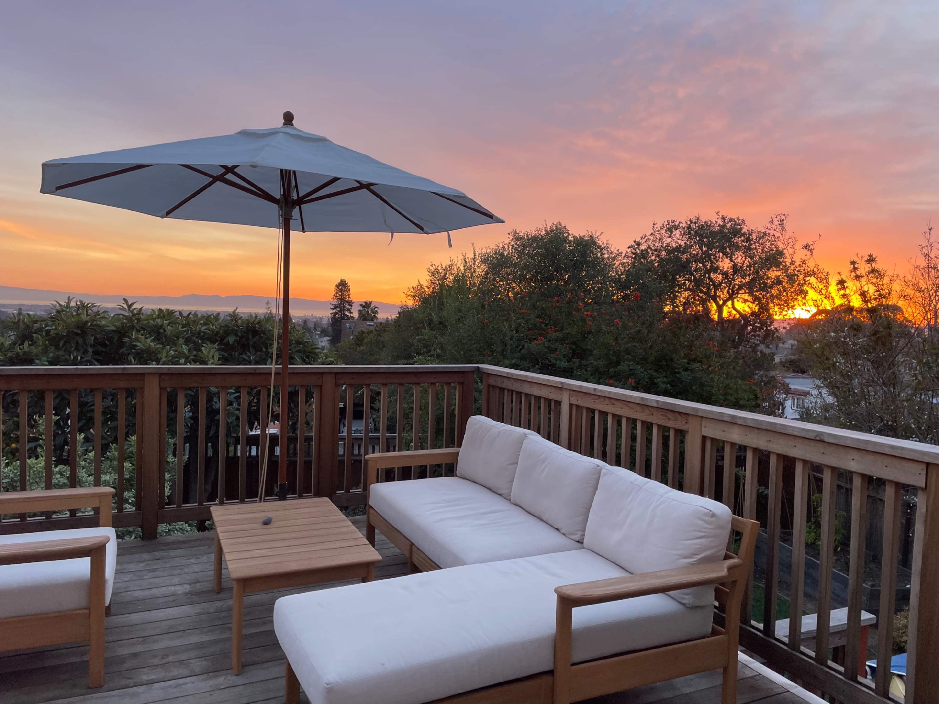 A wooden deck with a sofa and table is shaded by a large umbrella, overlooking a sunset with vibrant colors in the sky.