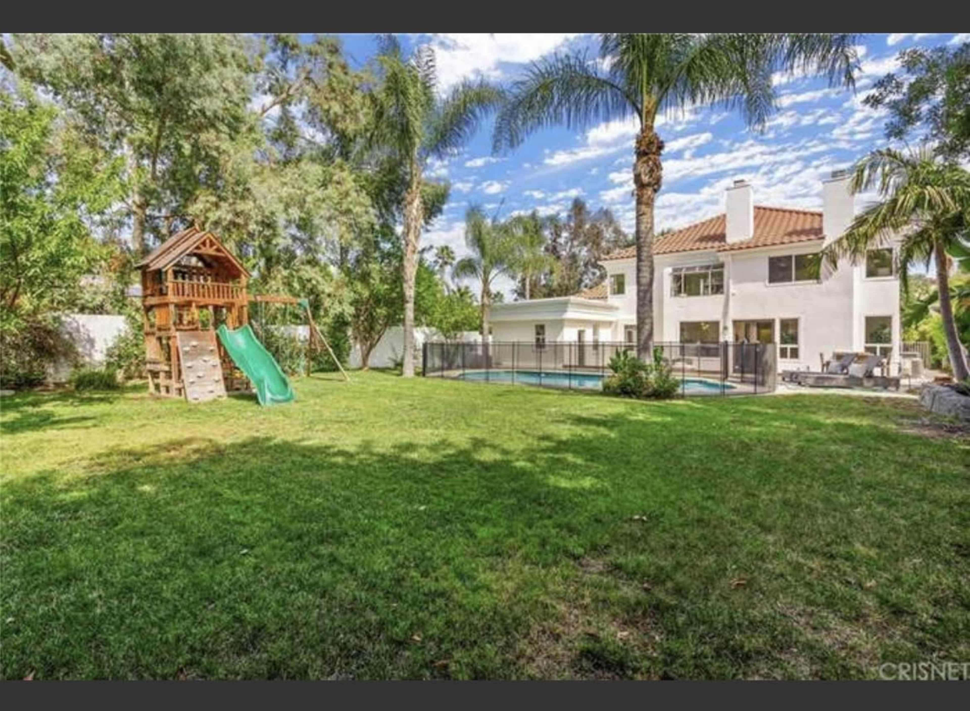 A backyard with a play structure featuring a slide, a swimming pool enclosed by a fence, and lush greenery surrounding a two-story house.