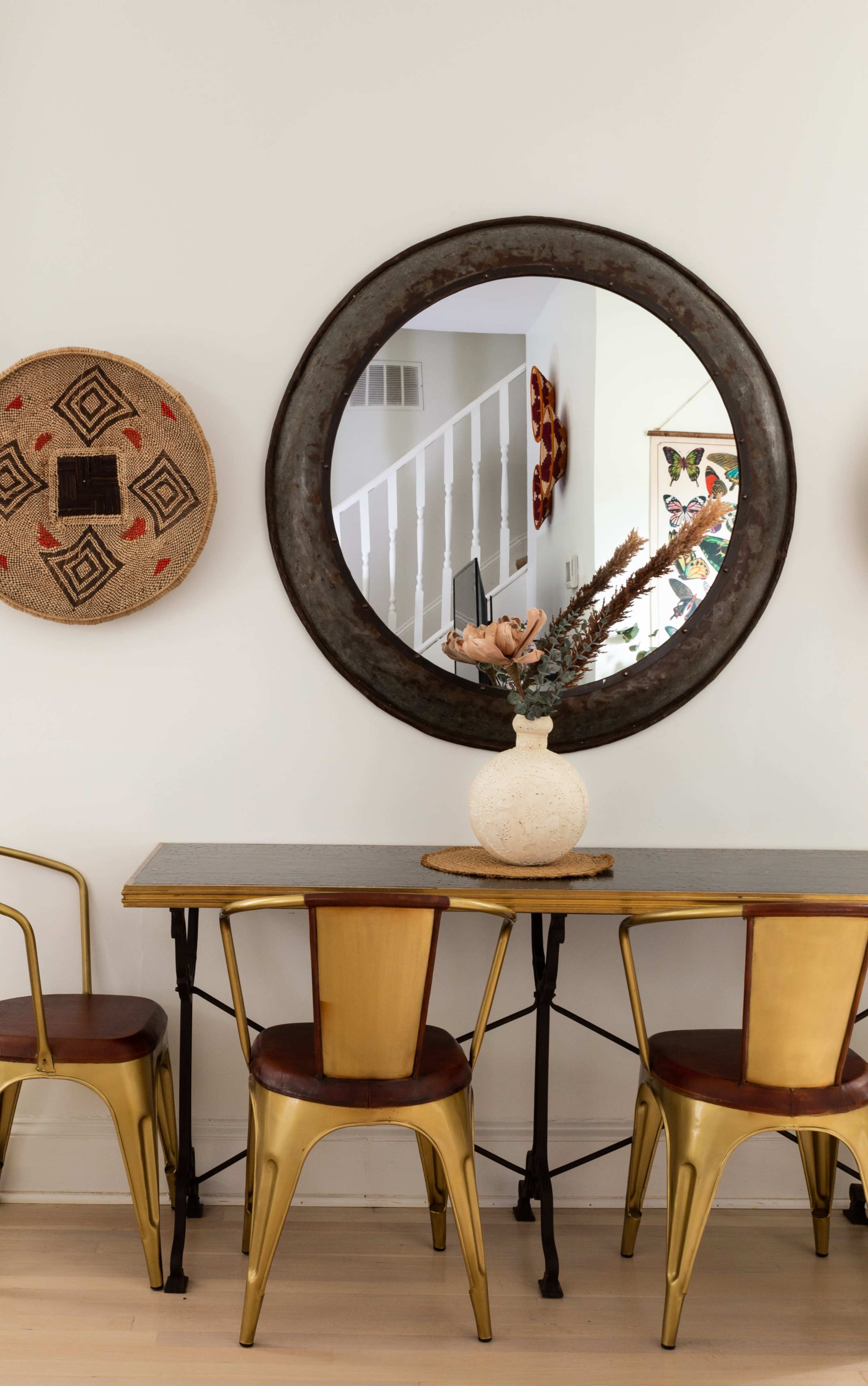 A circular mirror hangs above a table with metallic chairs, while woven wall art is displayed on either side.