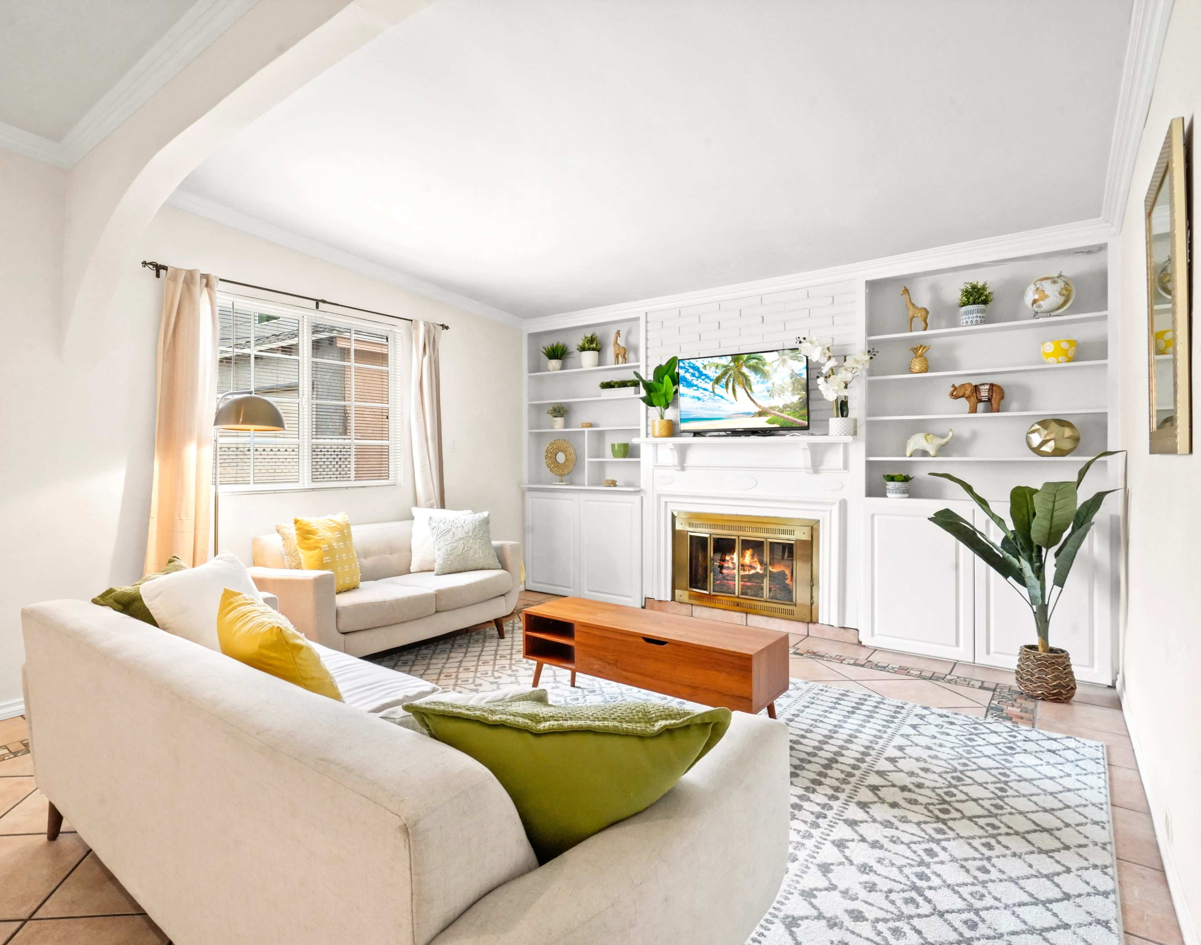 A living room featuring a light-colored sofa, a wooden coffee table, a TV mounted on a white shelving unit, and decorative items on the shelves.