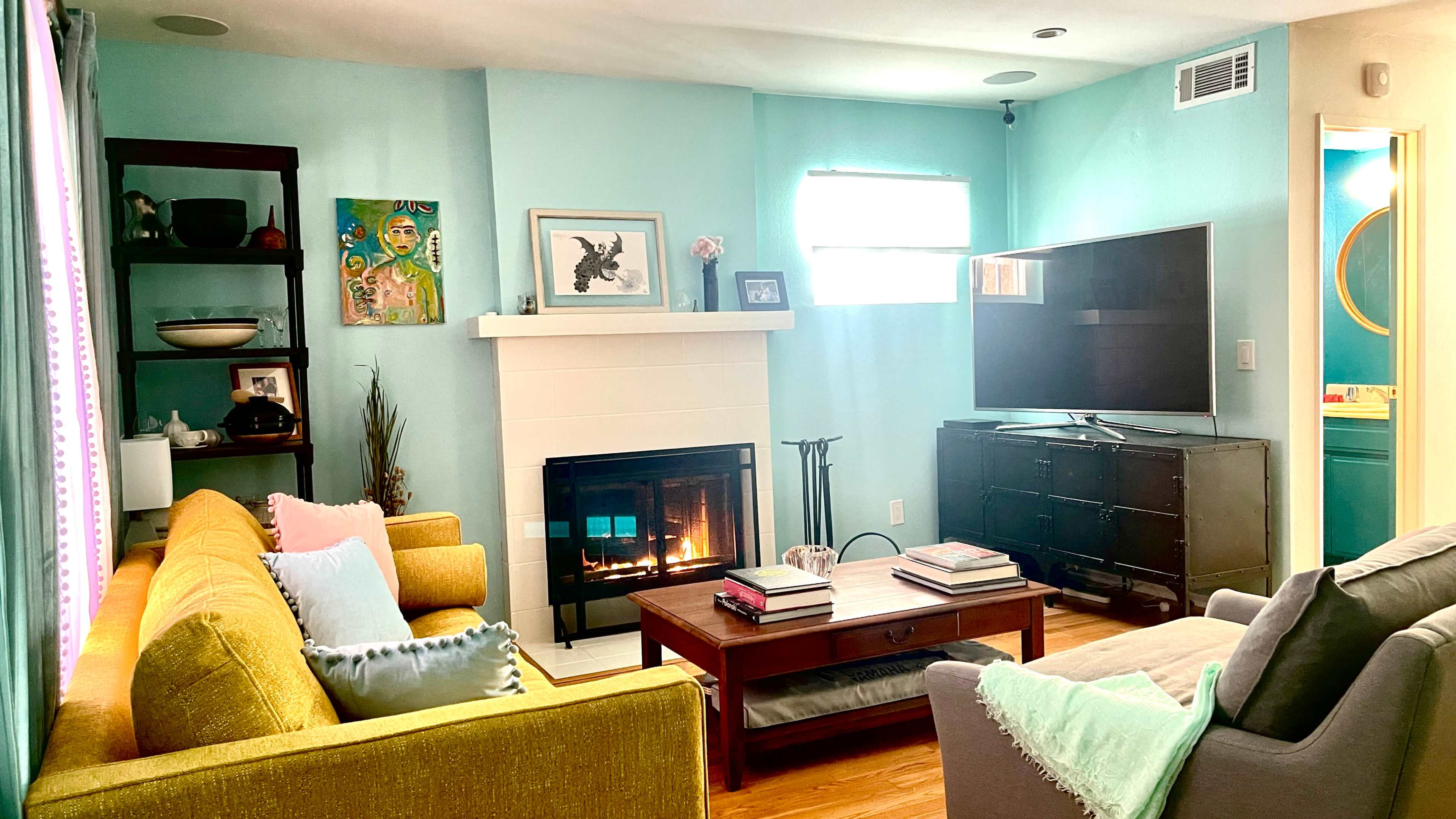 A brightly lit living room features a yellow sofa, a gray armchair, a fireplace, and a large TV on an entertainment unit.