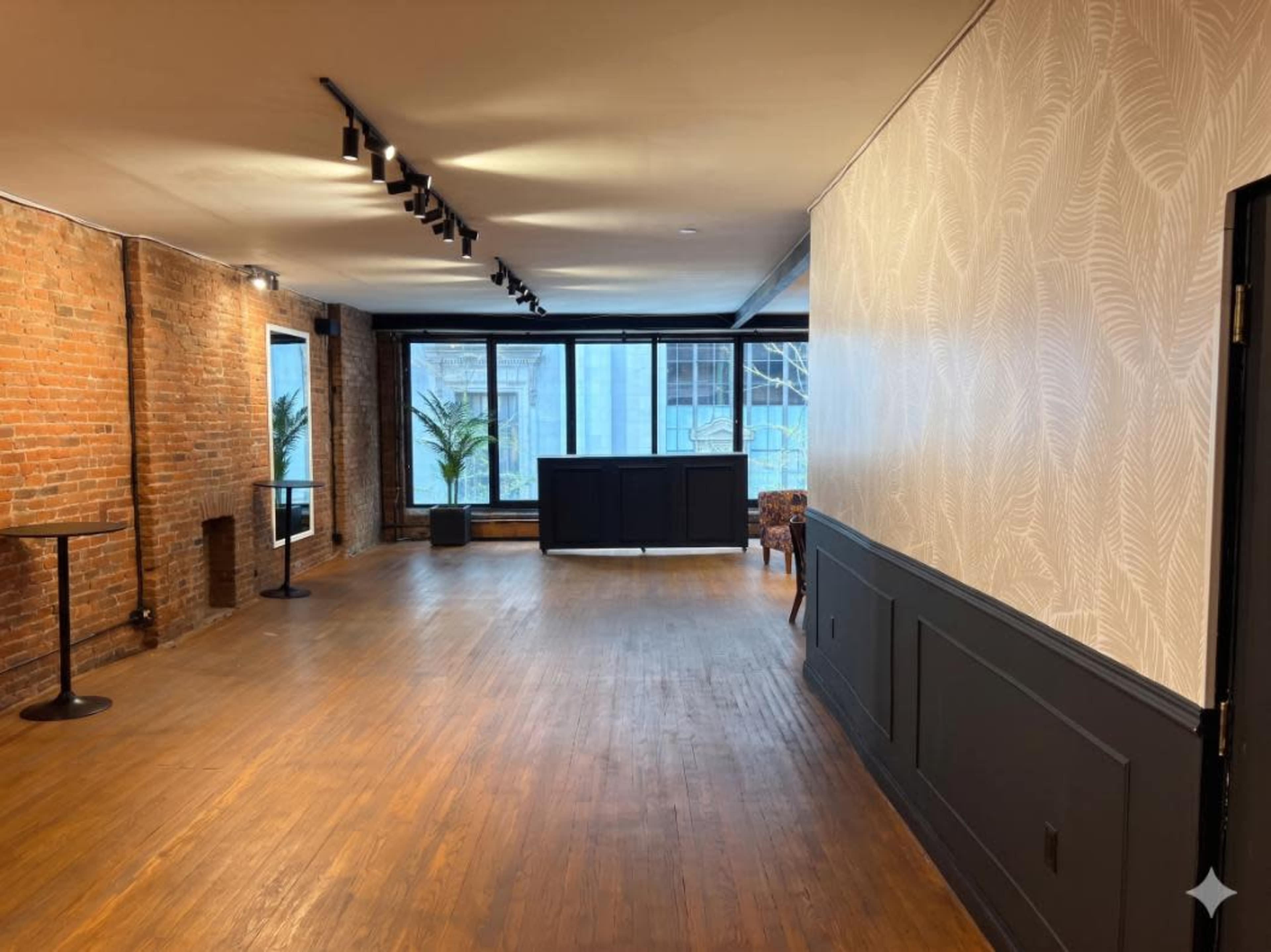 The image shows a spacious room with exposed brick walls, wooden flooring, and large windows that provide natural light, along with a small bar area and minimal furnishings.