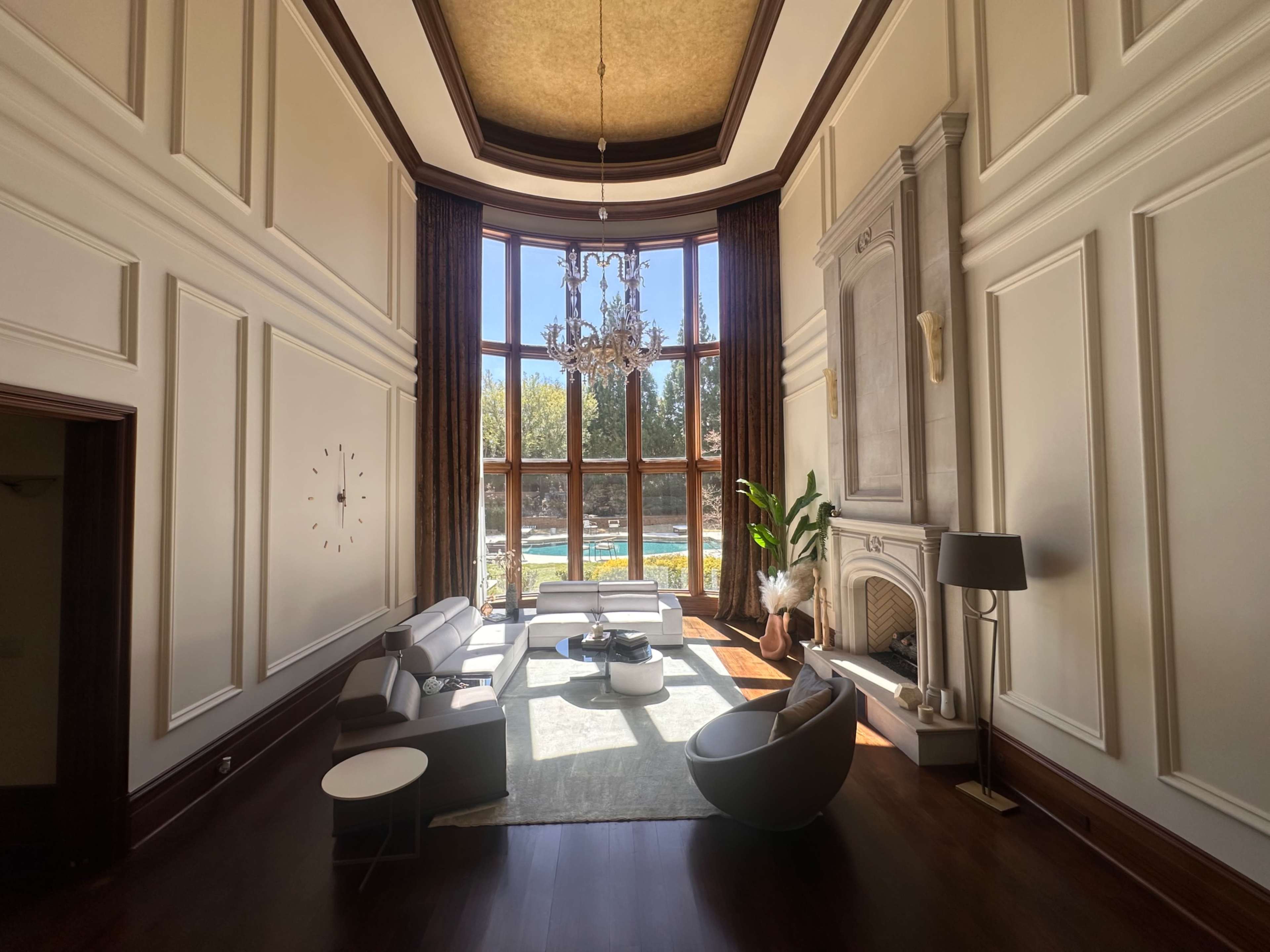 The image features a spacious living room with large windows overlooking a pool, adorned with a chandelier, modern furniture, and elegant decor.