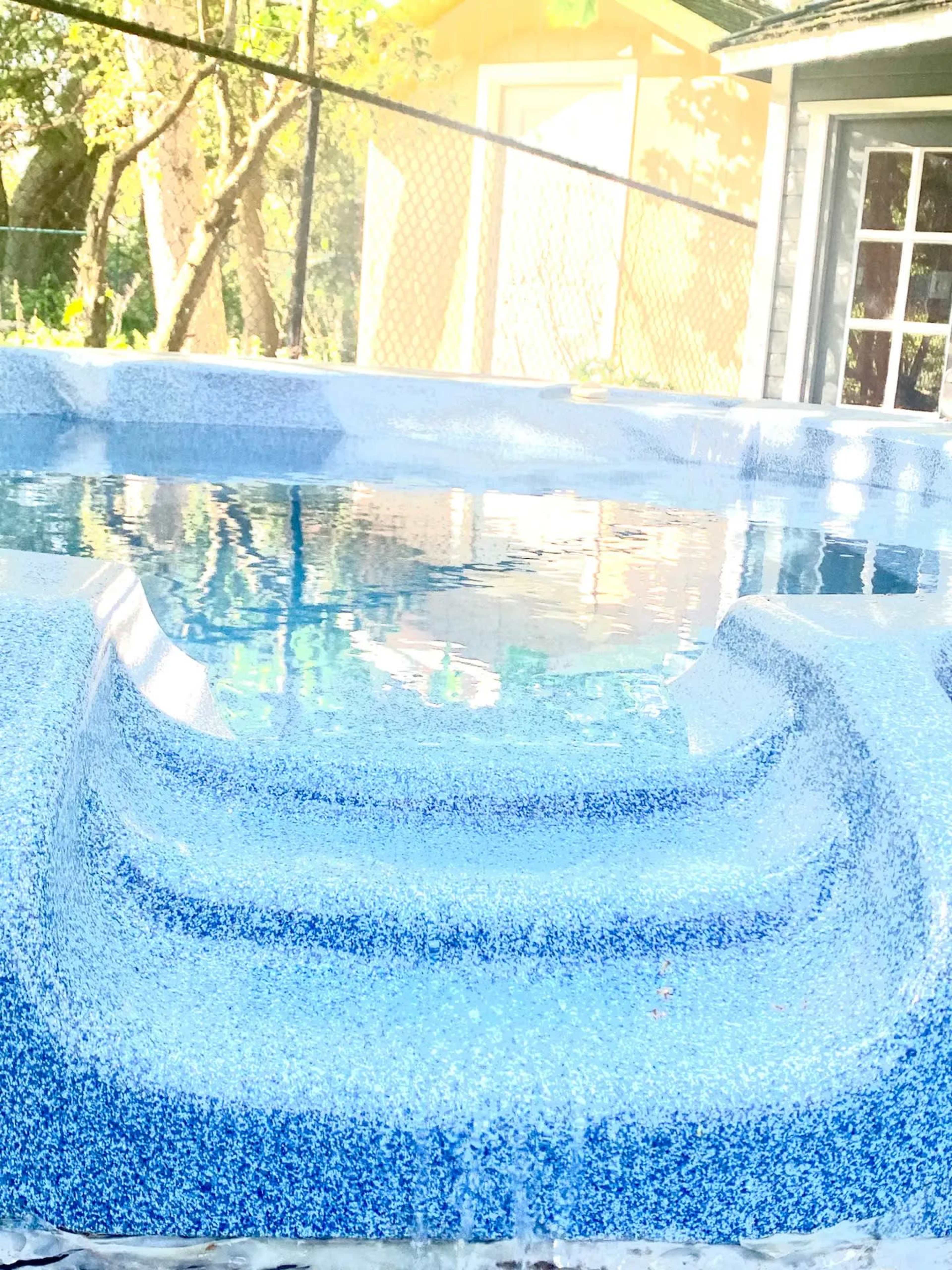 A partially filled swimming pool with textured steps leading into the water, surrounded by trees and a house in the background.