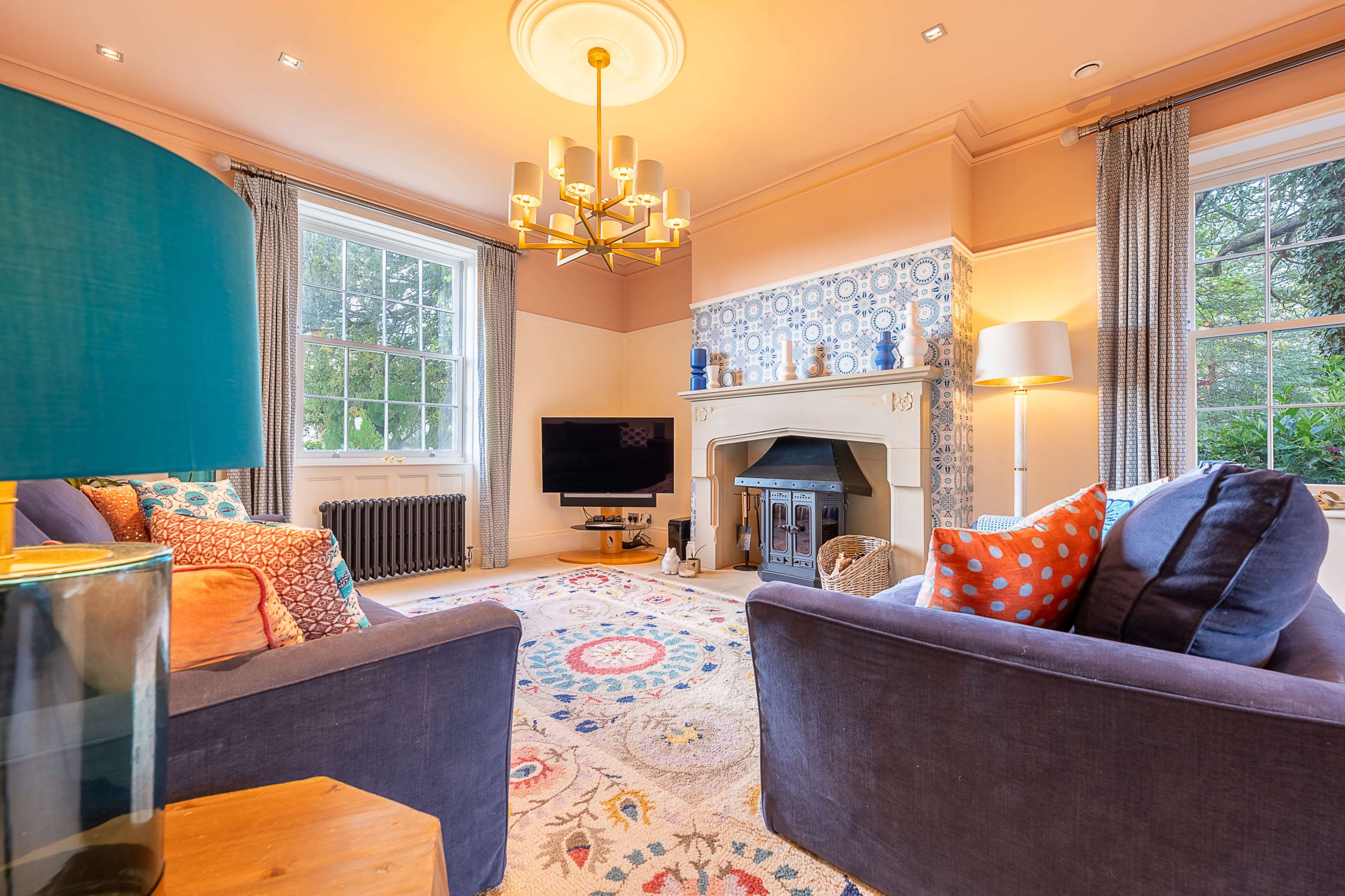 A cozy living room features a traditional fireplace, patterned rug, modern television, and colorful furnishings around a large window.