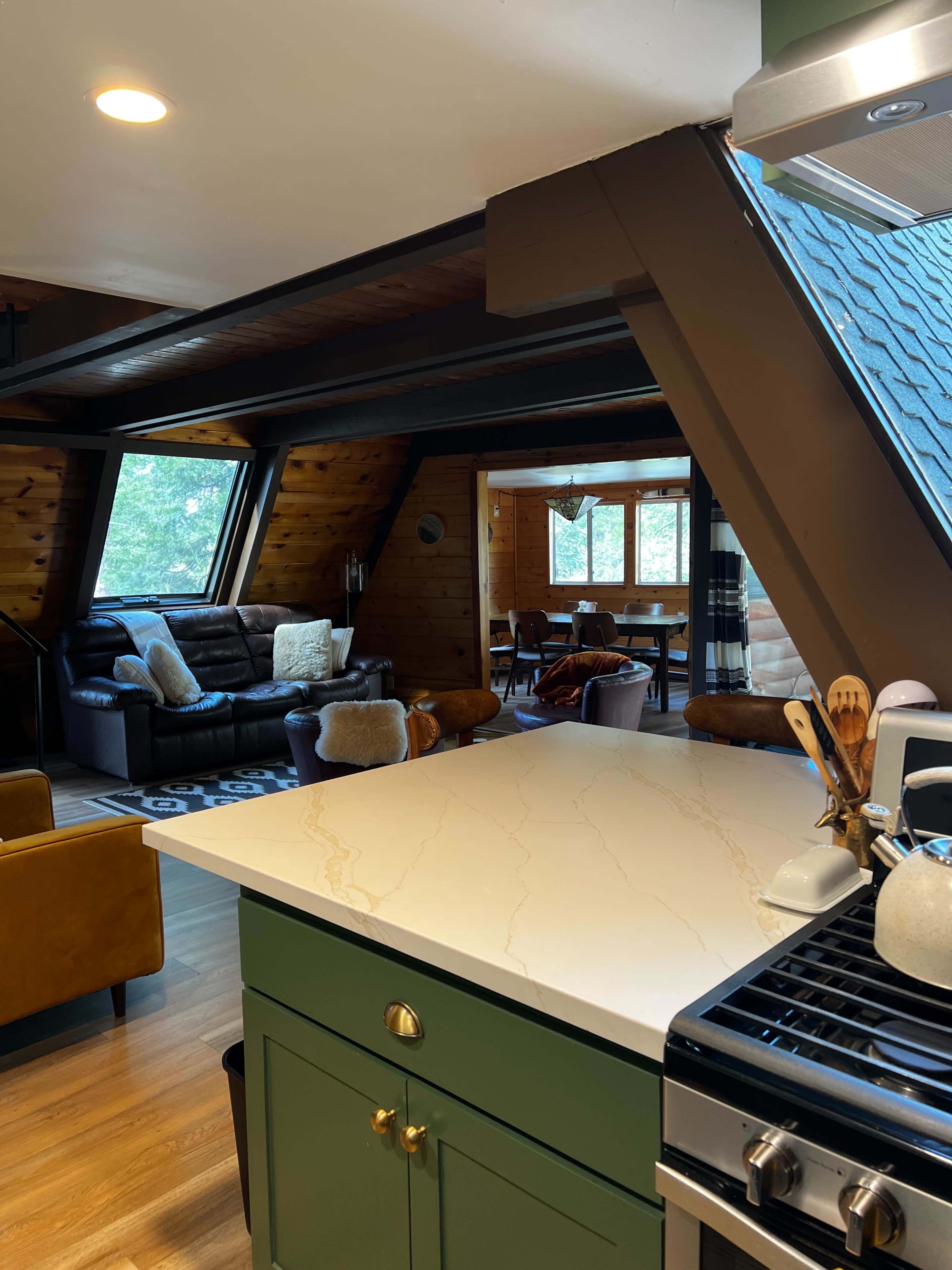 A modern kitchen with a green island and a gas stove, overlooking a living area with leather sofas and a dining space in a cozy cabin setting.