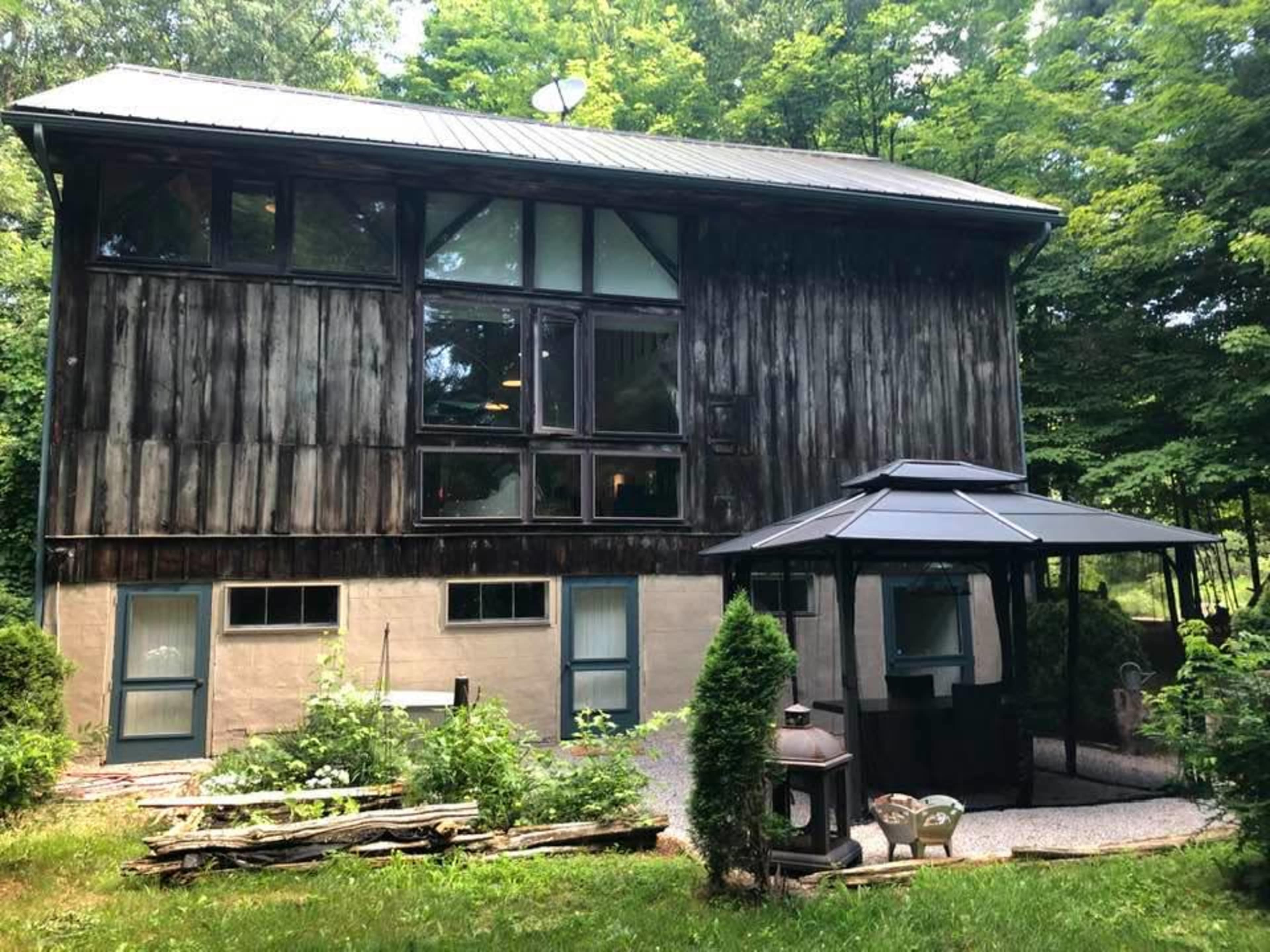A two-story wooden house surrounded by trees, with a gazebo and a gravel pathway in the foreground.