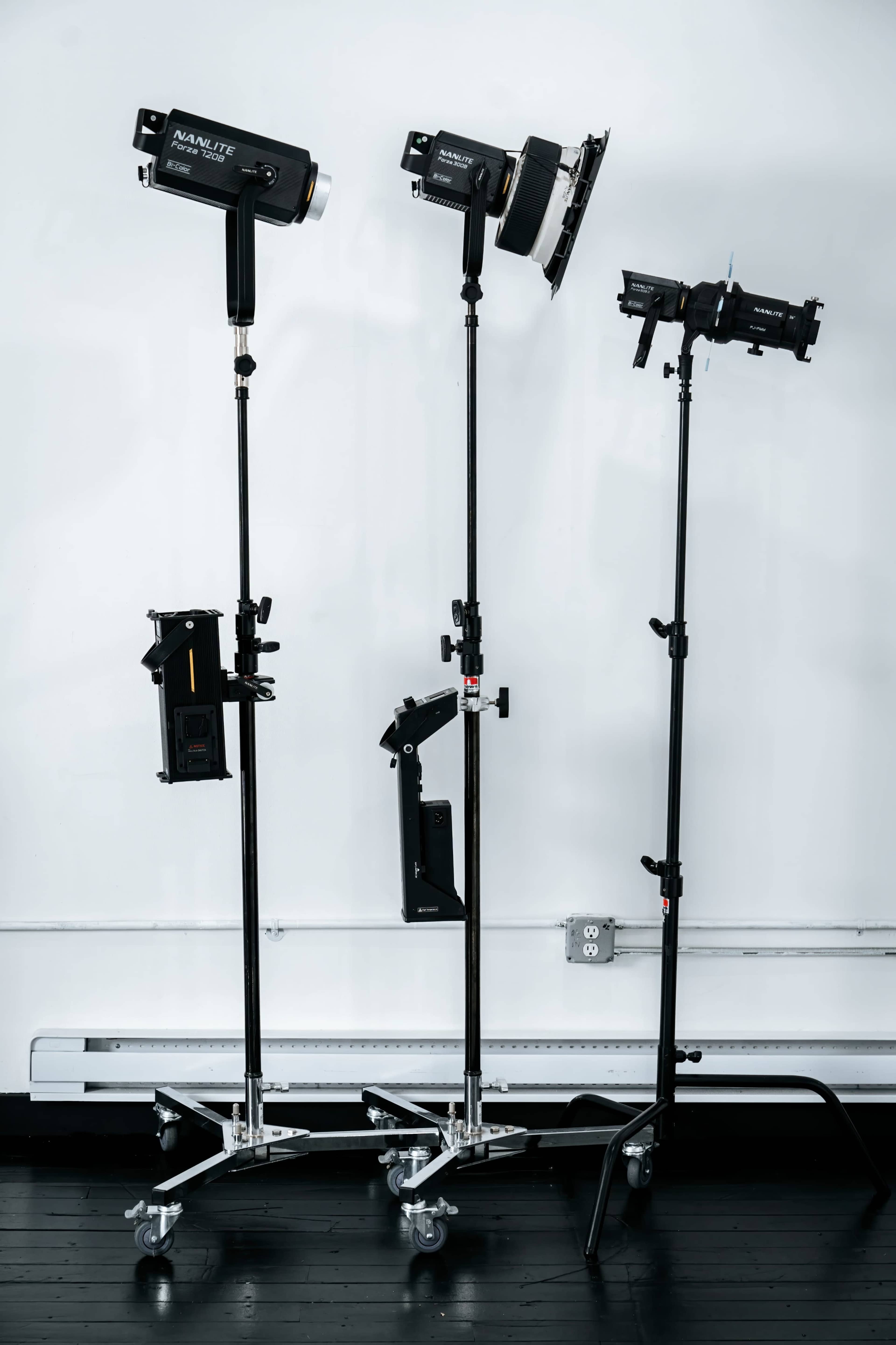 Three studio lights mounted on adjustable stands in an interior space with a plain wall.
