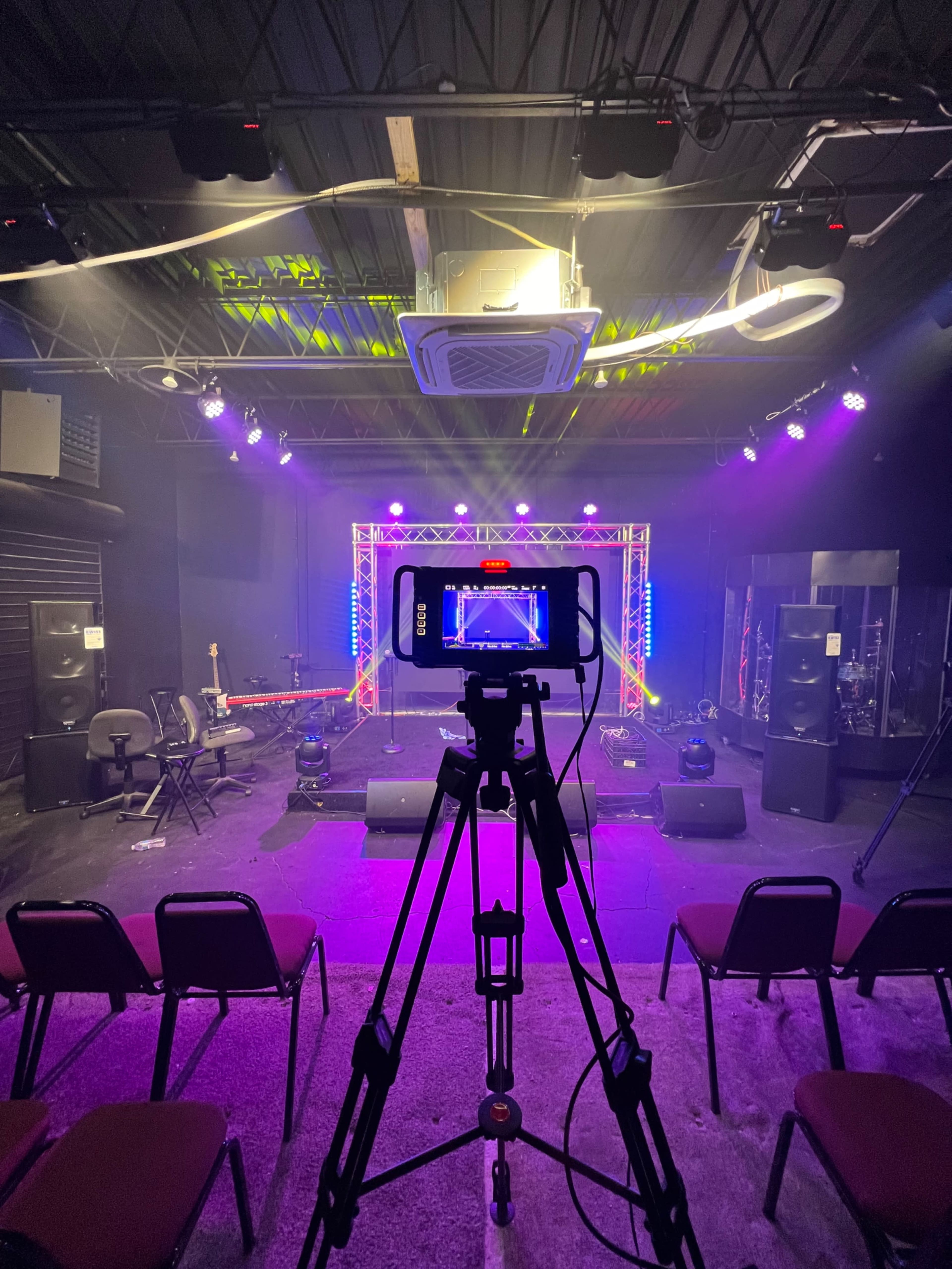 A camera is positioned in front of a stage illuminated by colorful lights, with chairs set up for an audience.