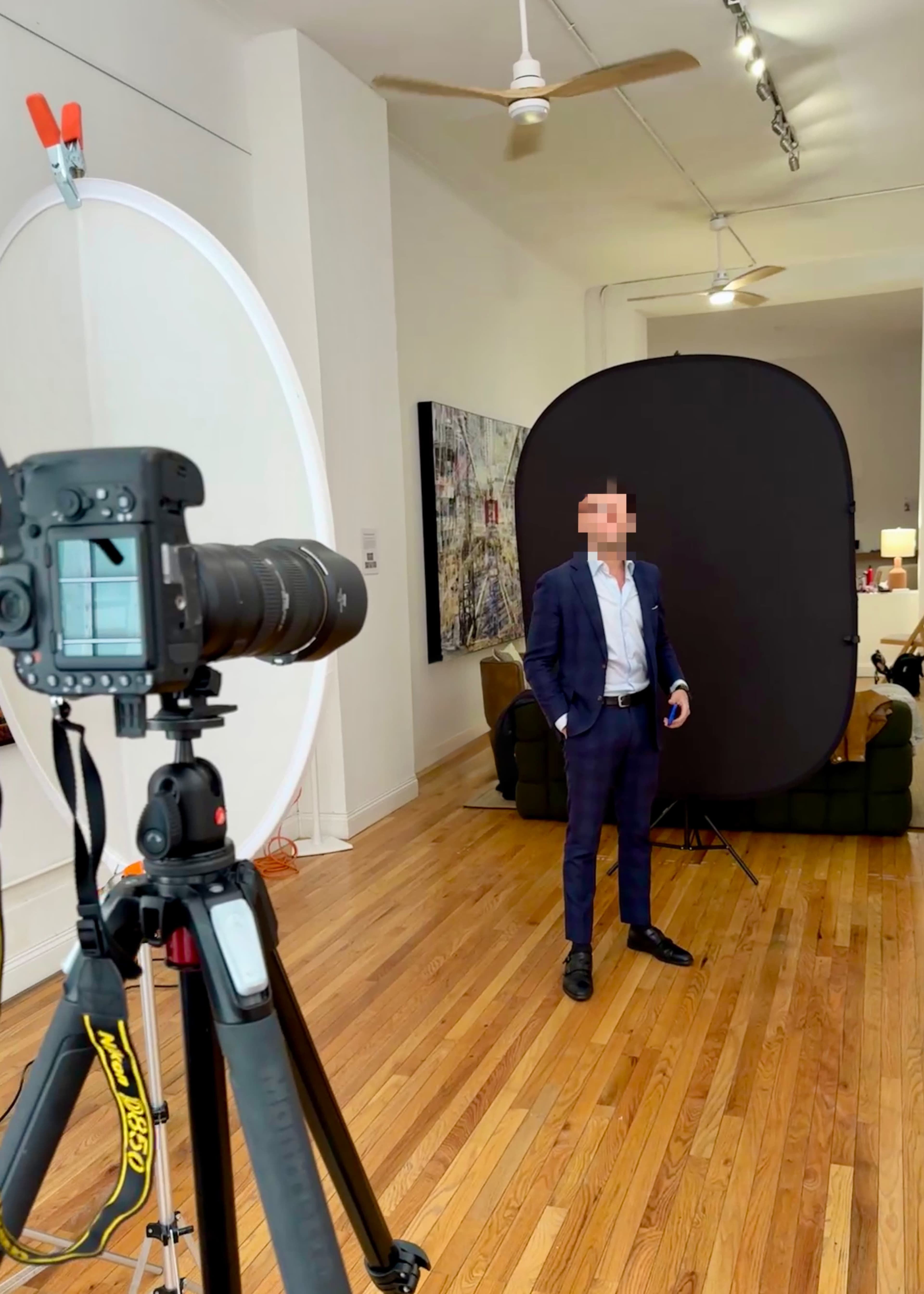 A man in a blue suit stands in front of a black backdrop while a camera on a tripod points towards him.