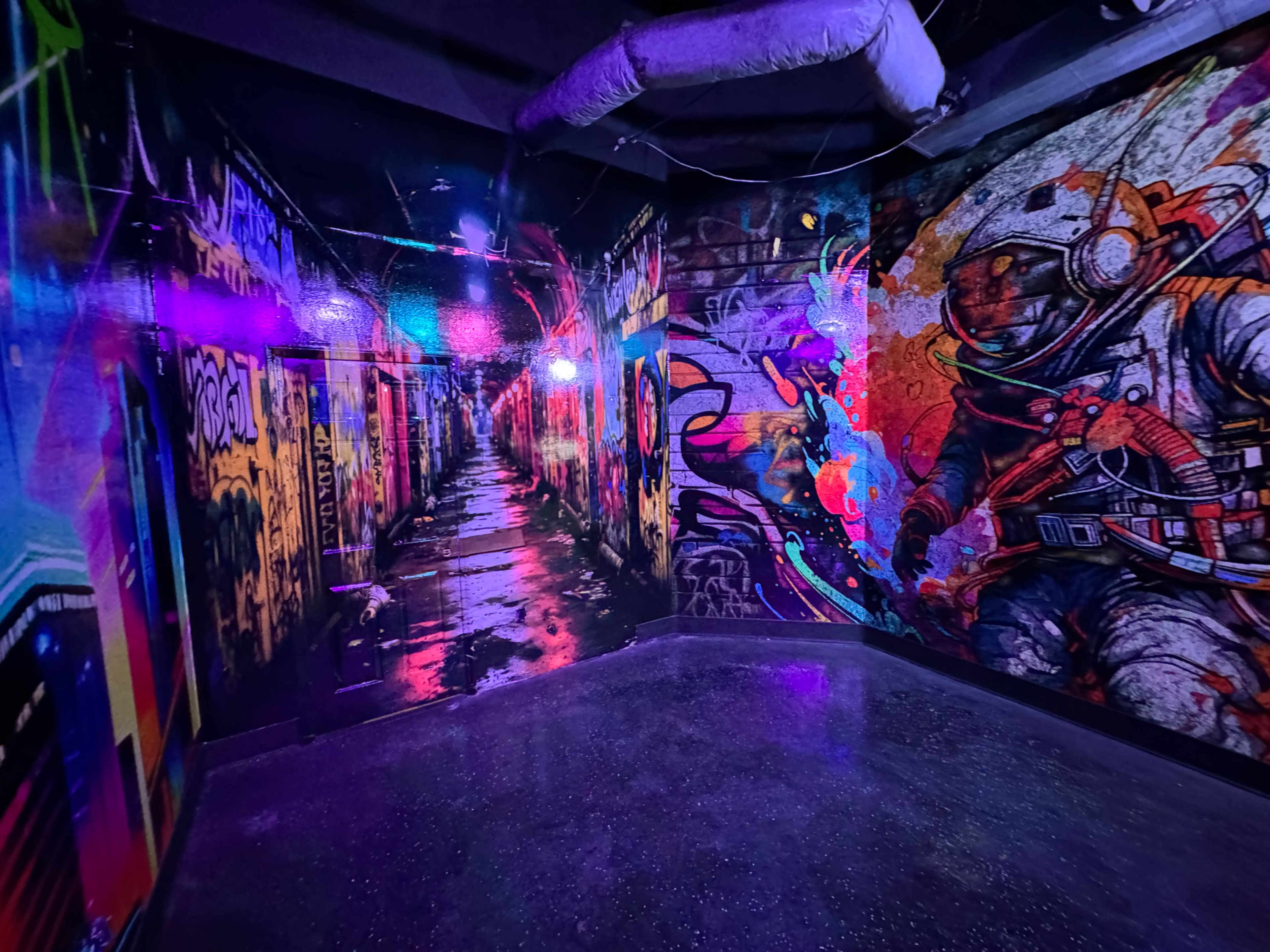The image shows a dimly lit alleyway featuring vibrant street art on the walls, with bright colors and a reflective surface creating a dynamic atmosphere.